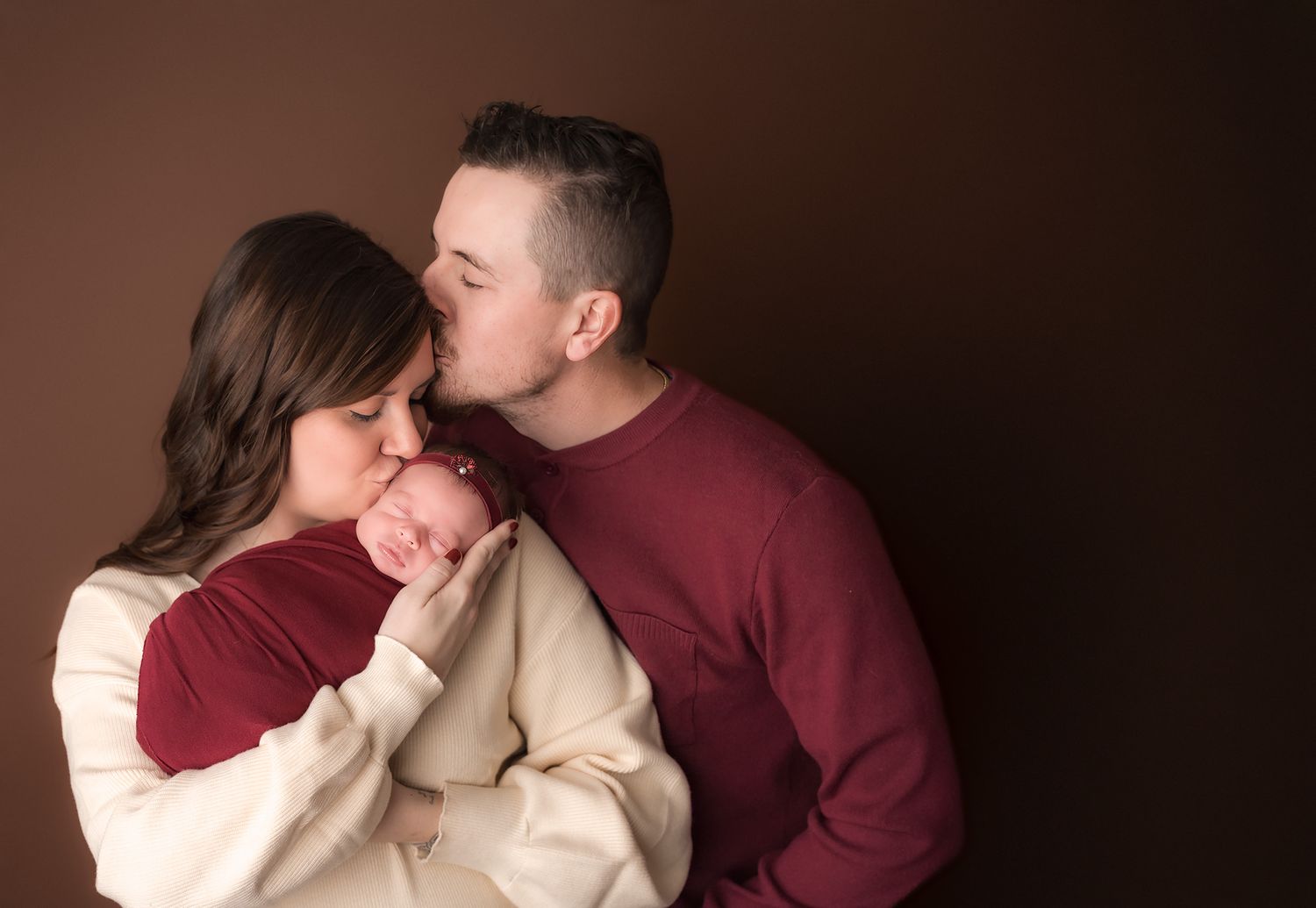 New parents share a tender moment, father kissing mother's head as she cradles their sleeping newborn baby.