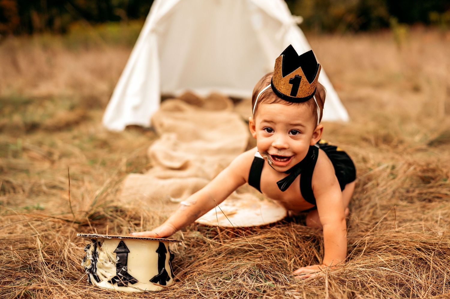 Smiling toddler in black outfit and crown crawls on hay near a white teepee during outdoor cake smash photo session.