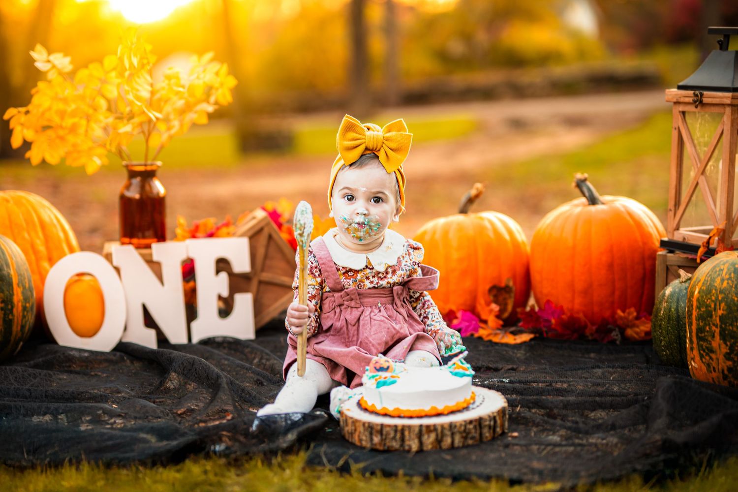Baby girl in pink dress celebrates first birthday with pumpkins and ONE sign in golden autumn outdoor setting.