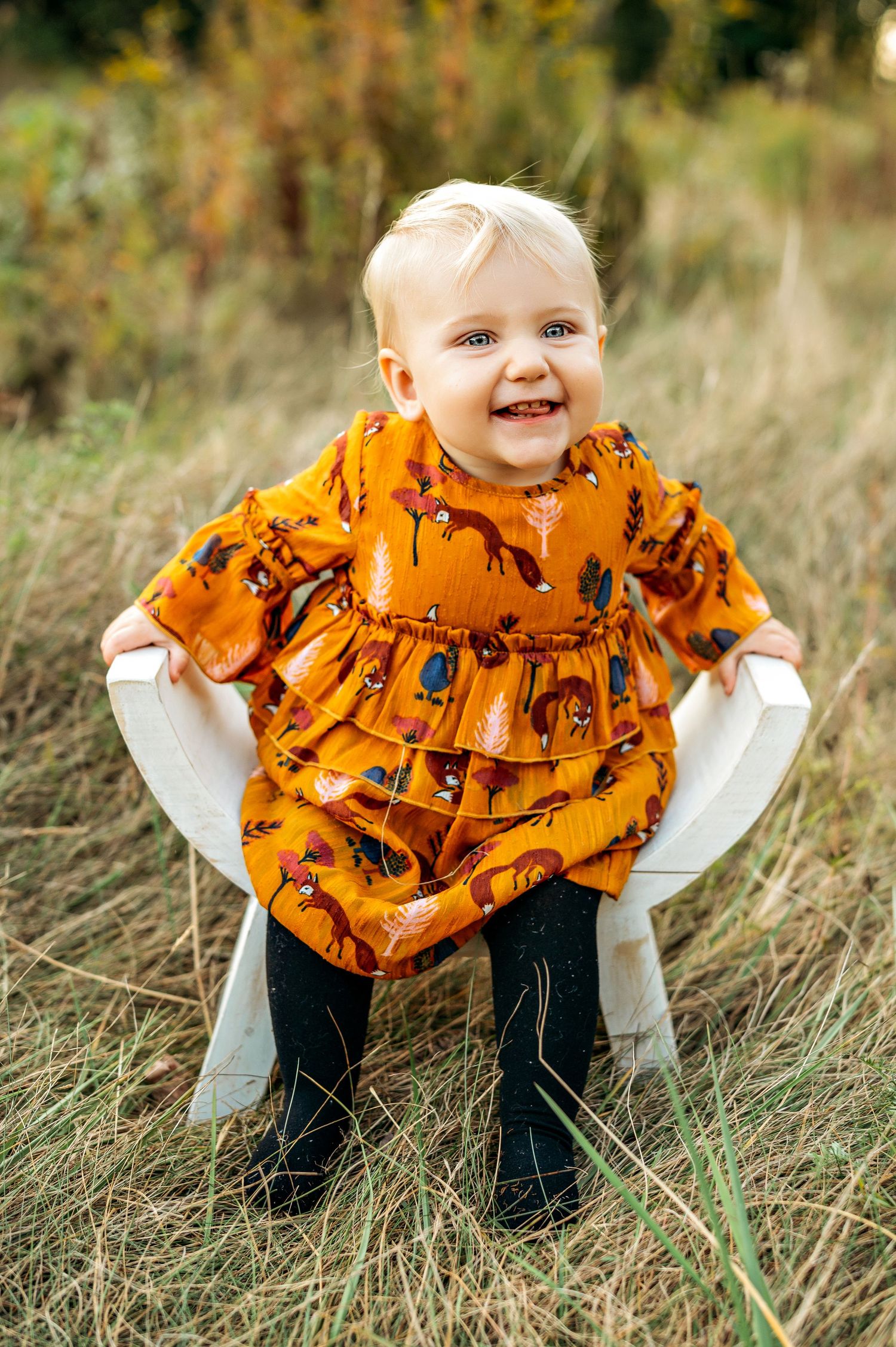 Smiling toddler girl in orange fox-print dress sitting on white chair in autumn field.