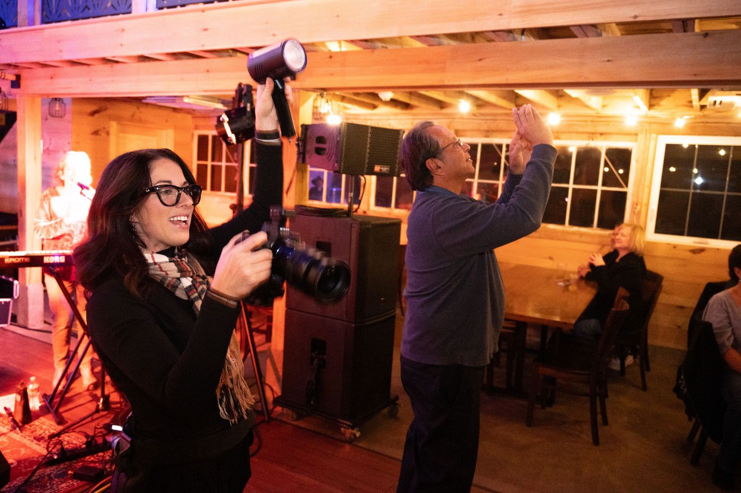 Two photographers capturing moments at an indoor evening event with string lights and warm ambient lighting.