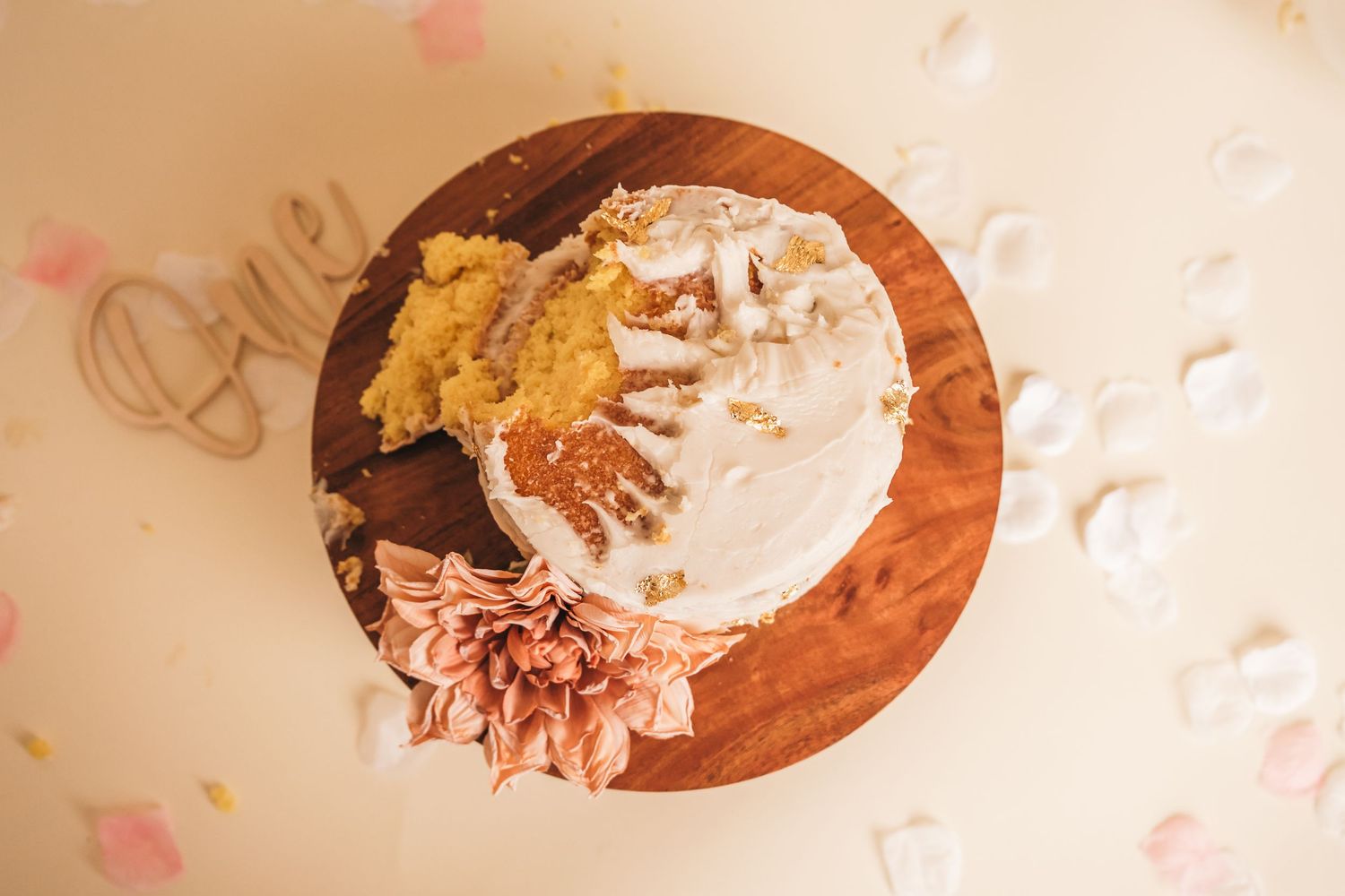 Overhead view of a smash cake on a wooden board with white frosting, gold accents, and a pink flower decoration.