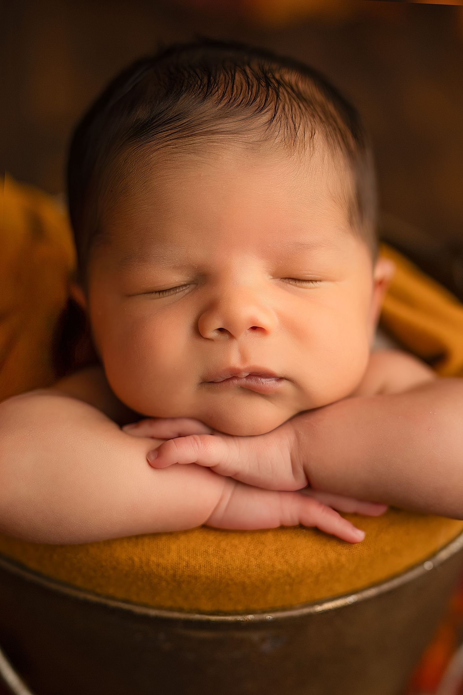 Peaceful newborn baby sleeping with hands folded under chin in a golden bowl prop.