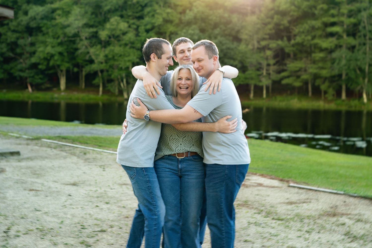 Family of four hugging and smiling together on a sandy shore with a lake and green trees in background.