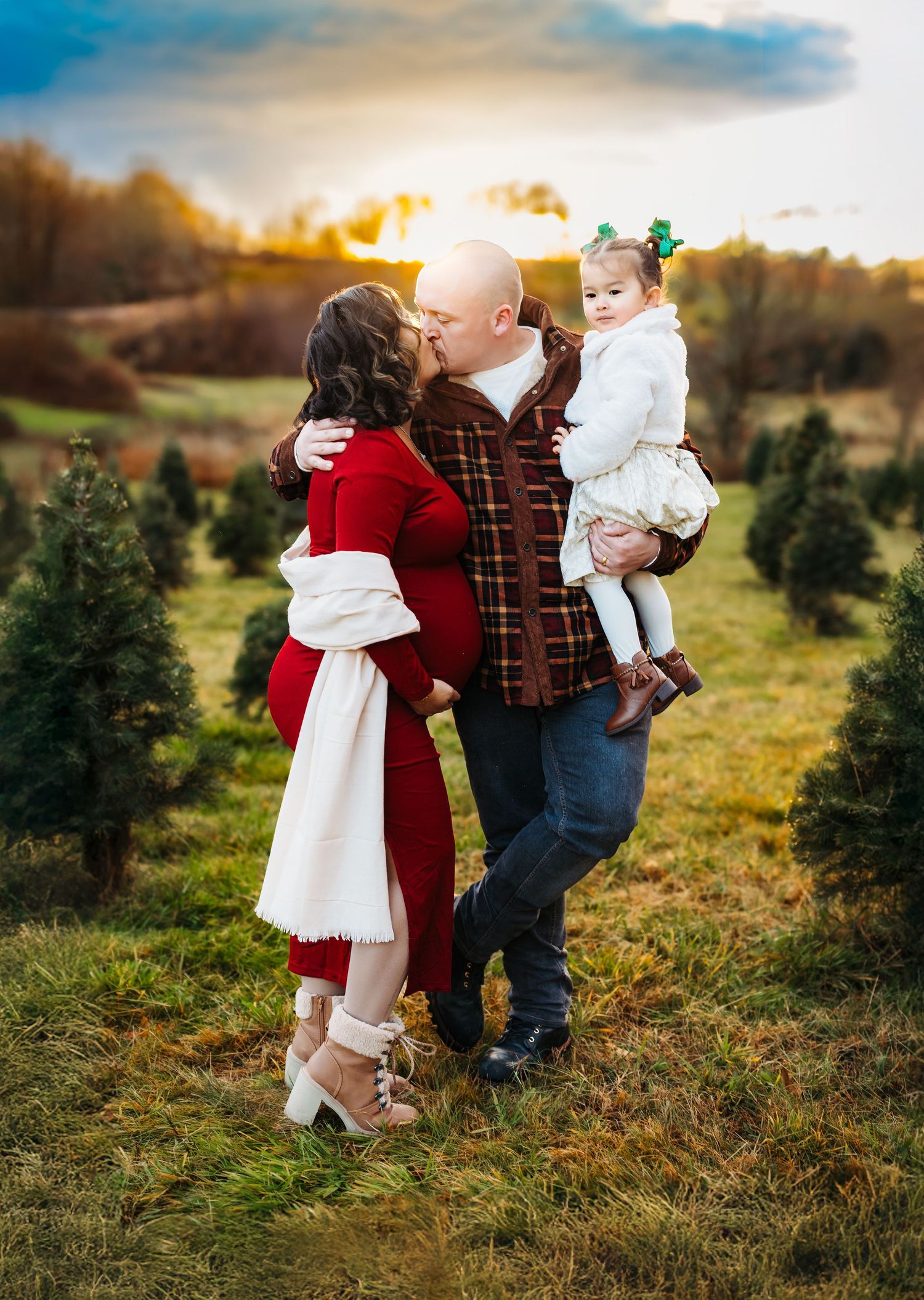 Family Christmas tree farm photo with couple kissing, man holding toddler girl, wearing red and plaid outfits at sunset.