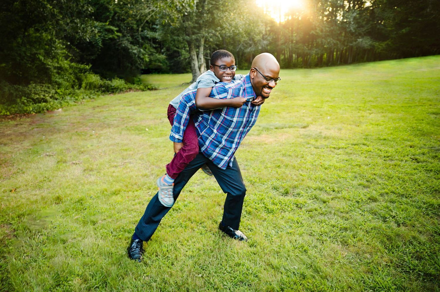 A father gives his laughing child a piggyback ride on a sunny green lawn with trees in the background.