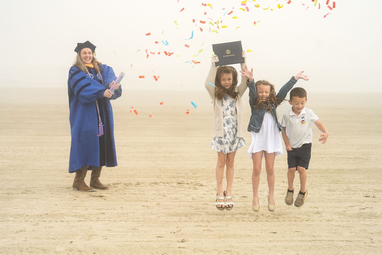 Graduate in blue cap and gown celebrates with three children tossing colorful confetti on a misty beach.