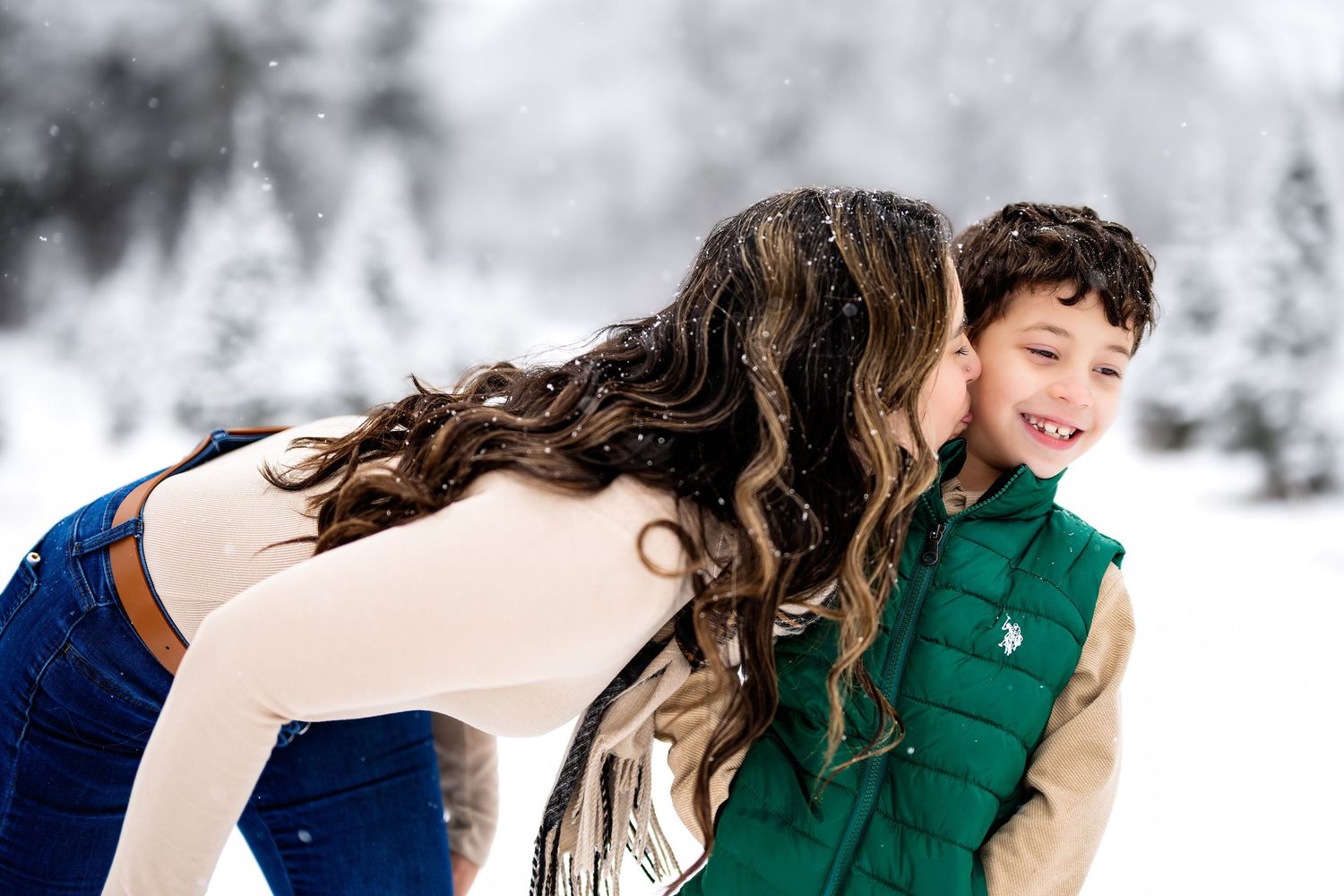 A mother kisses her young son on the cheek outdoors in a snowy winter wonderland setting.