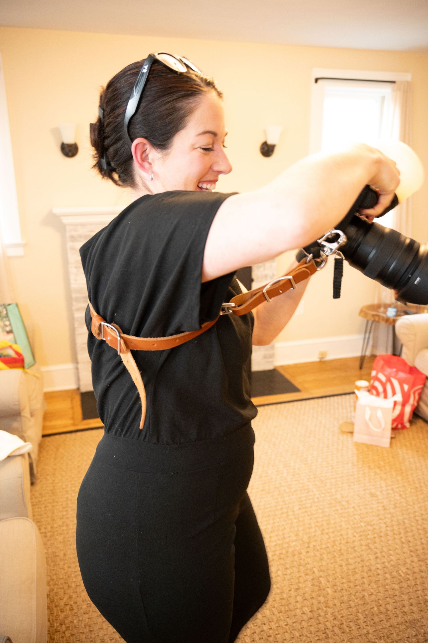 Woman in black outfit smiling while holding a camera with a brown leather strap indoors near a fireplace.