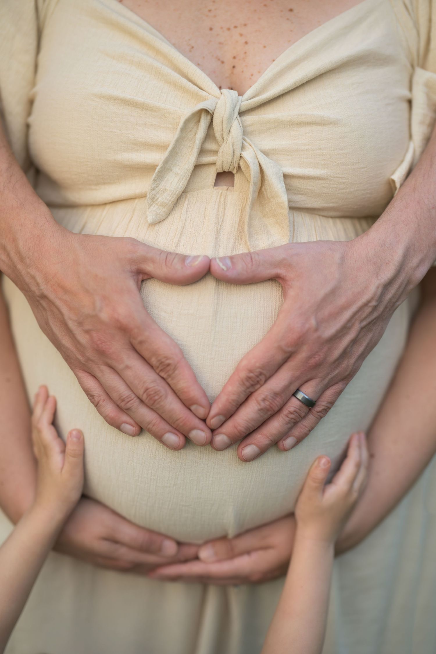 Pregnant woman in beige dress with partner and child forming heart shape with hands on baby bump.