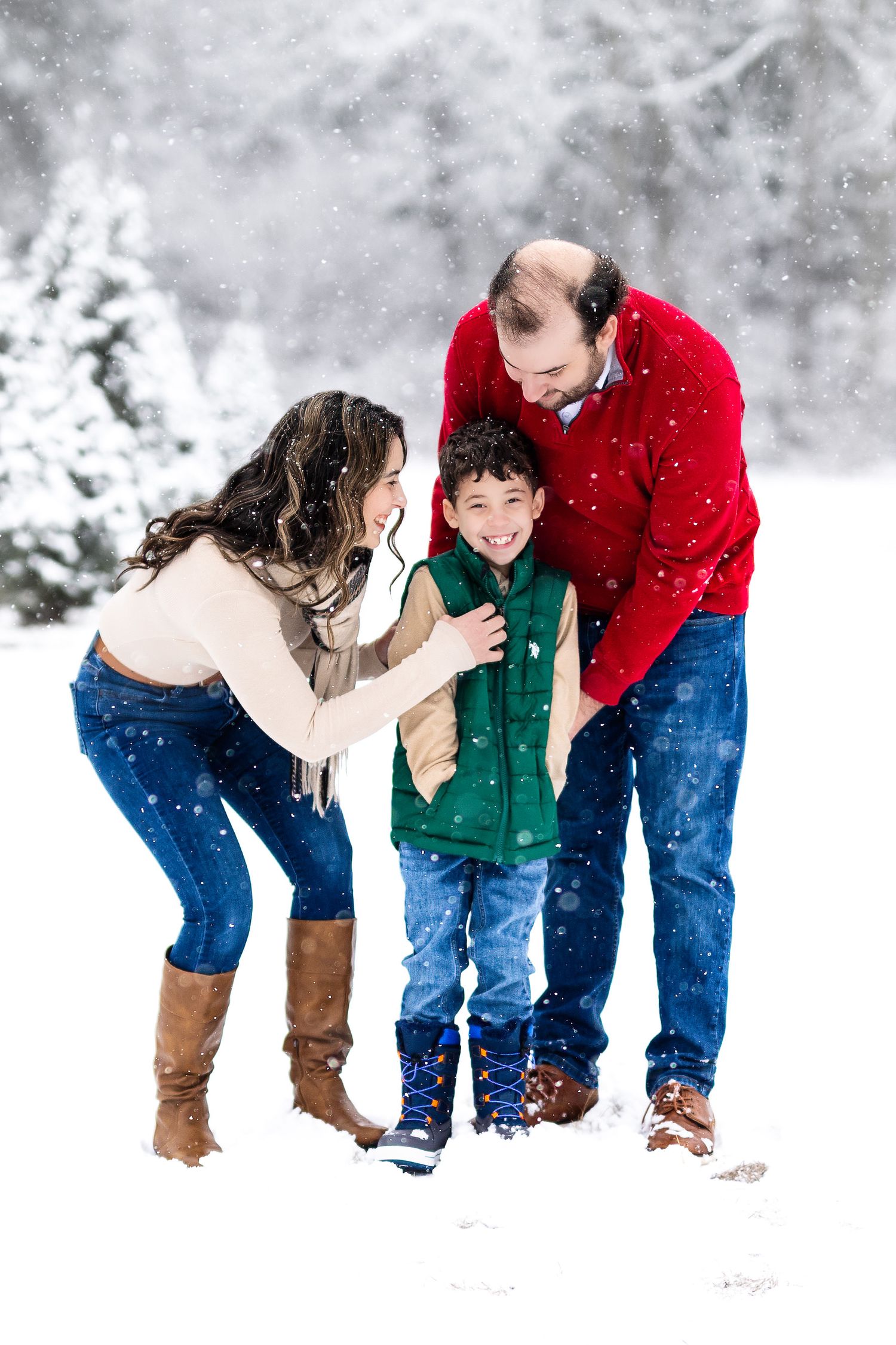 Happy family of three playing together in snowy winter landscape, parents in colorful sweaters with young son.