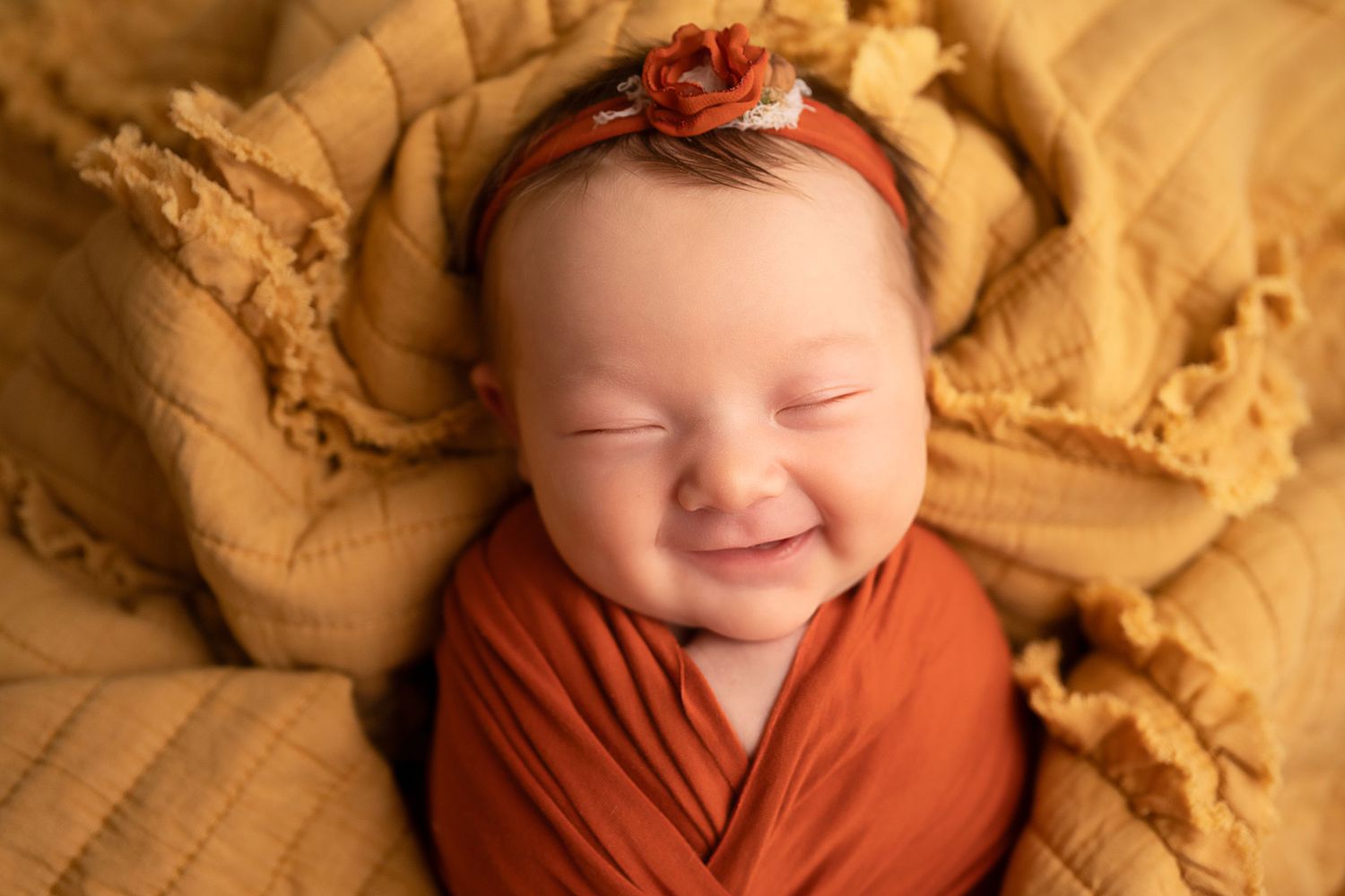Smiling newborn baby girl swaddled in rust orange wrap with floral headband on golden yellow textured blanket.