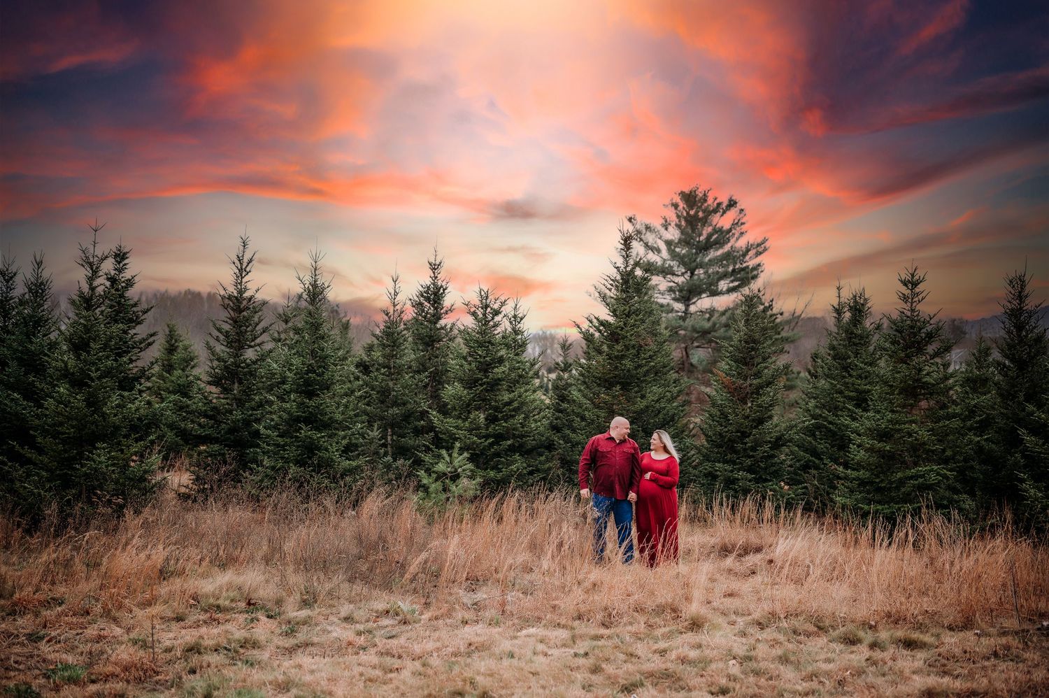 Couple in red walking through a Christmas tree farm at sunset with a dramatic colorful sky.