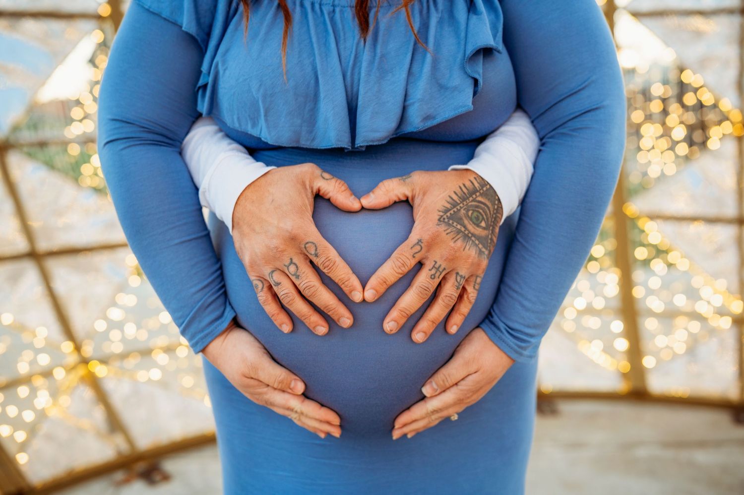 Pregnant woman in blue dress forming heart shape on baby bump with tattooed hands, bokeh lights background.