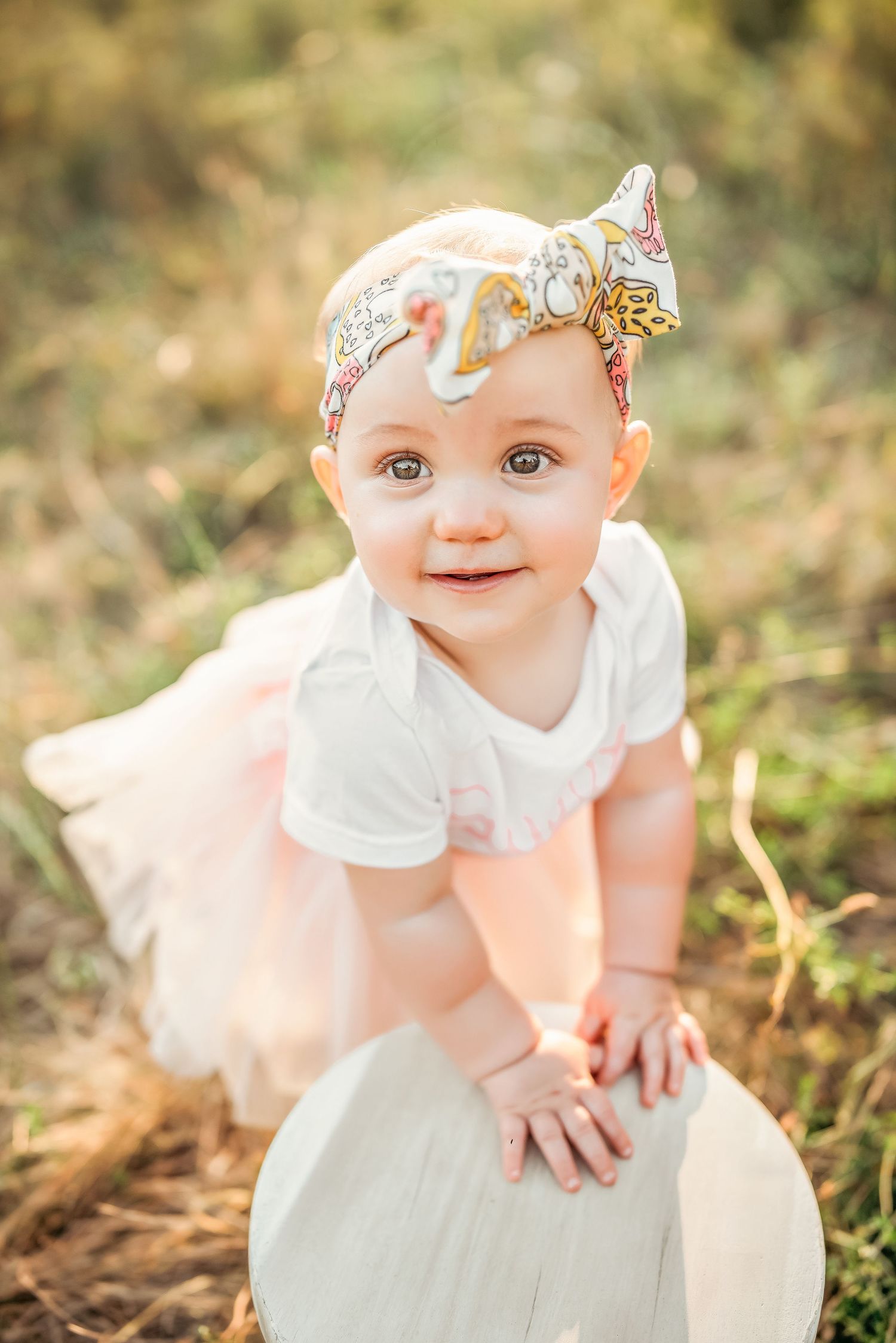 Adorable baby girl in white onesie and pink tutu with floral headband smiling outdoors in golden sunlight.