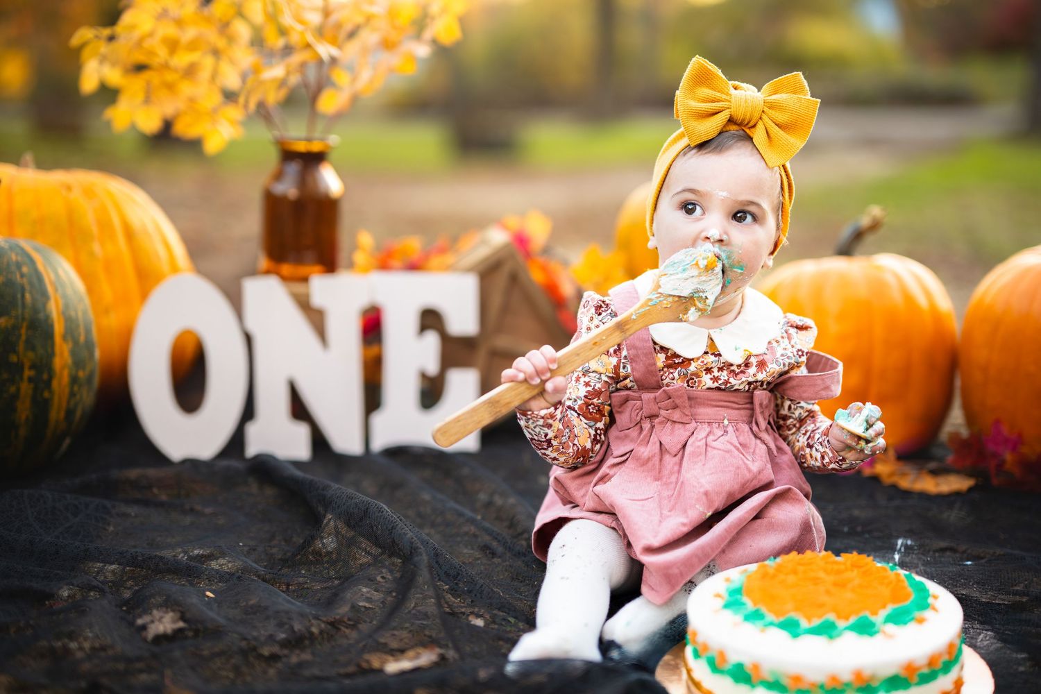 Baby girl in pink dress eating cake at fall first birthday party with pumpkins and ONE sign.