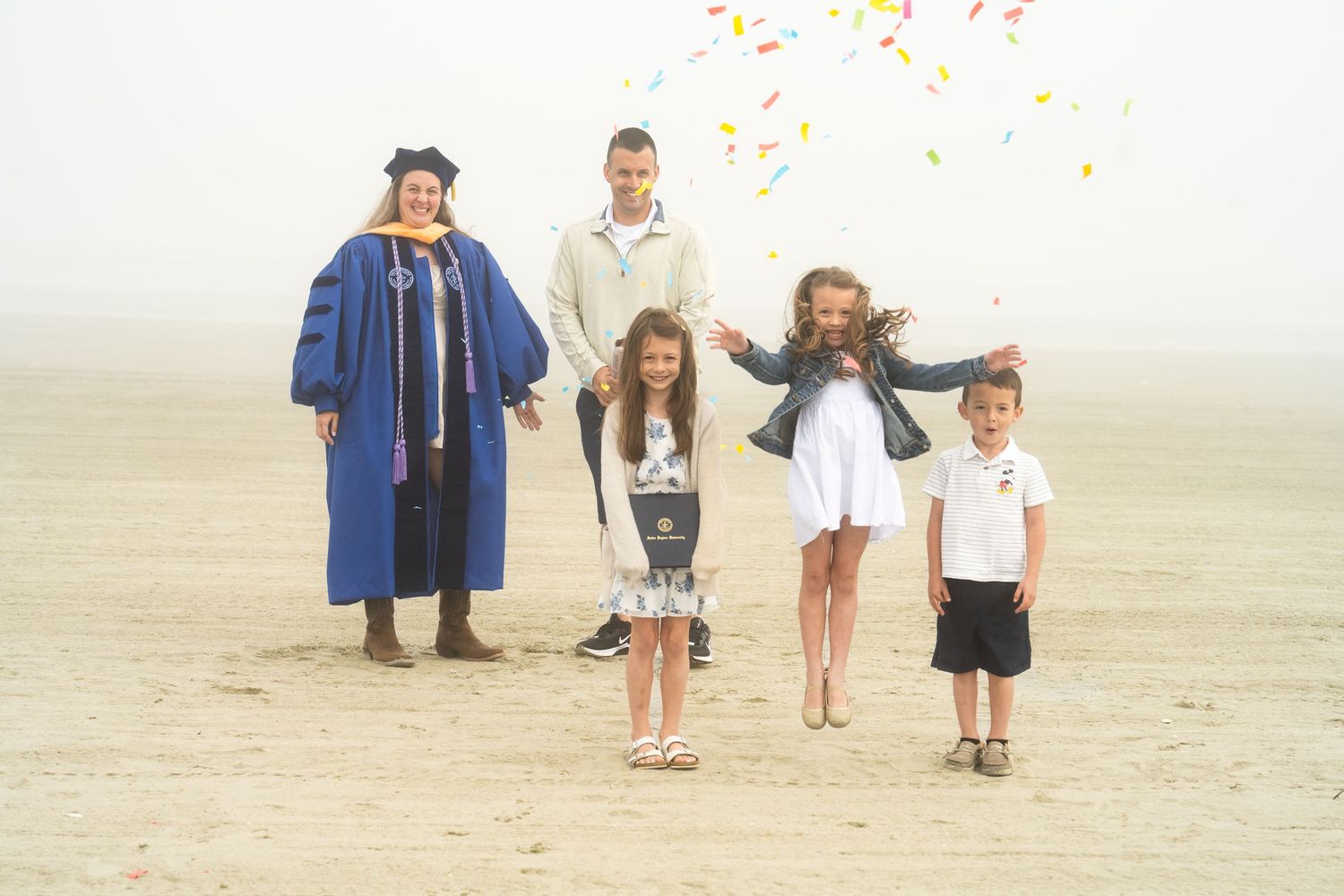Family celebrates graduation on a foggy beach, with woman in blue cap and gown surrounded by four children tossing confetti.