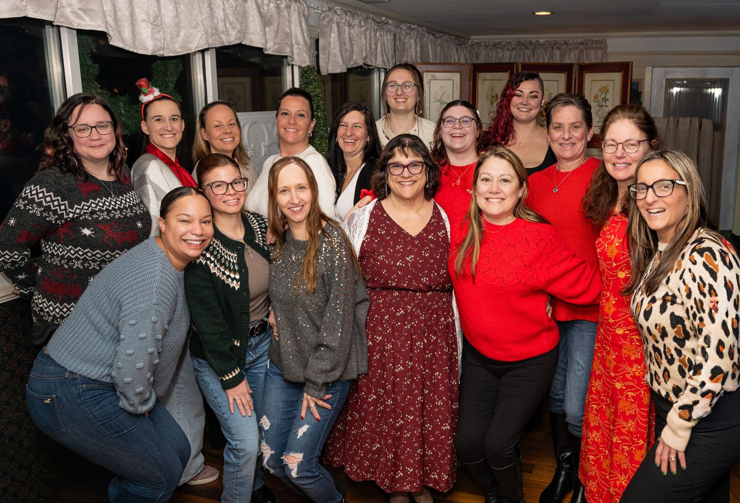 Group of women smiling together at a festive holiday party, wearing seasonal outfits in a decorated indoor venue.
