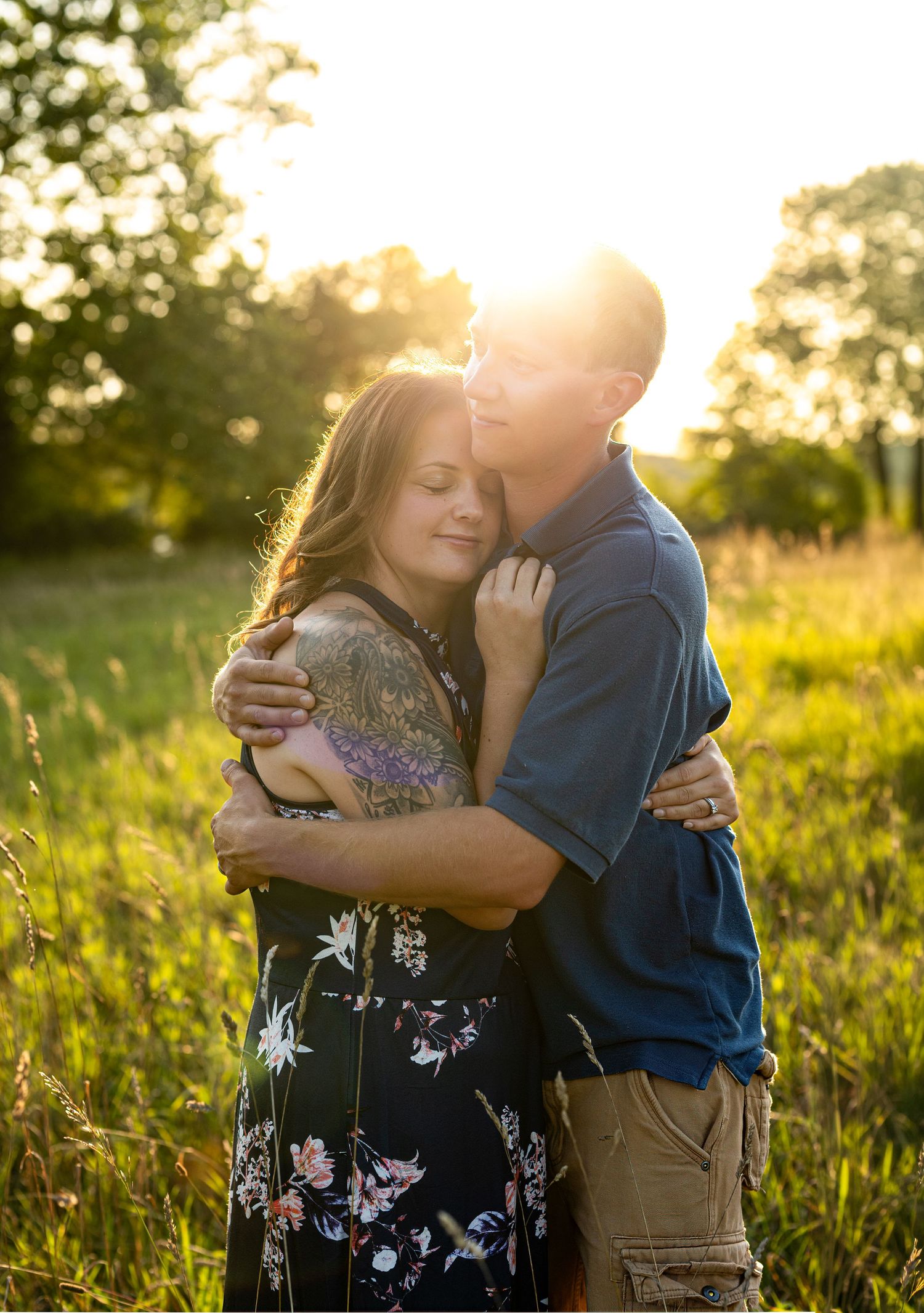 Couple embracing at golden hour in a sunlit meadow, woman with floral tattoo in a black floral dress.