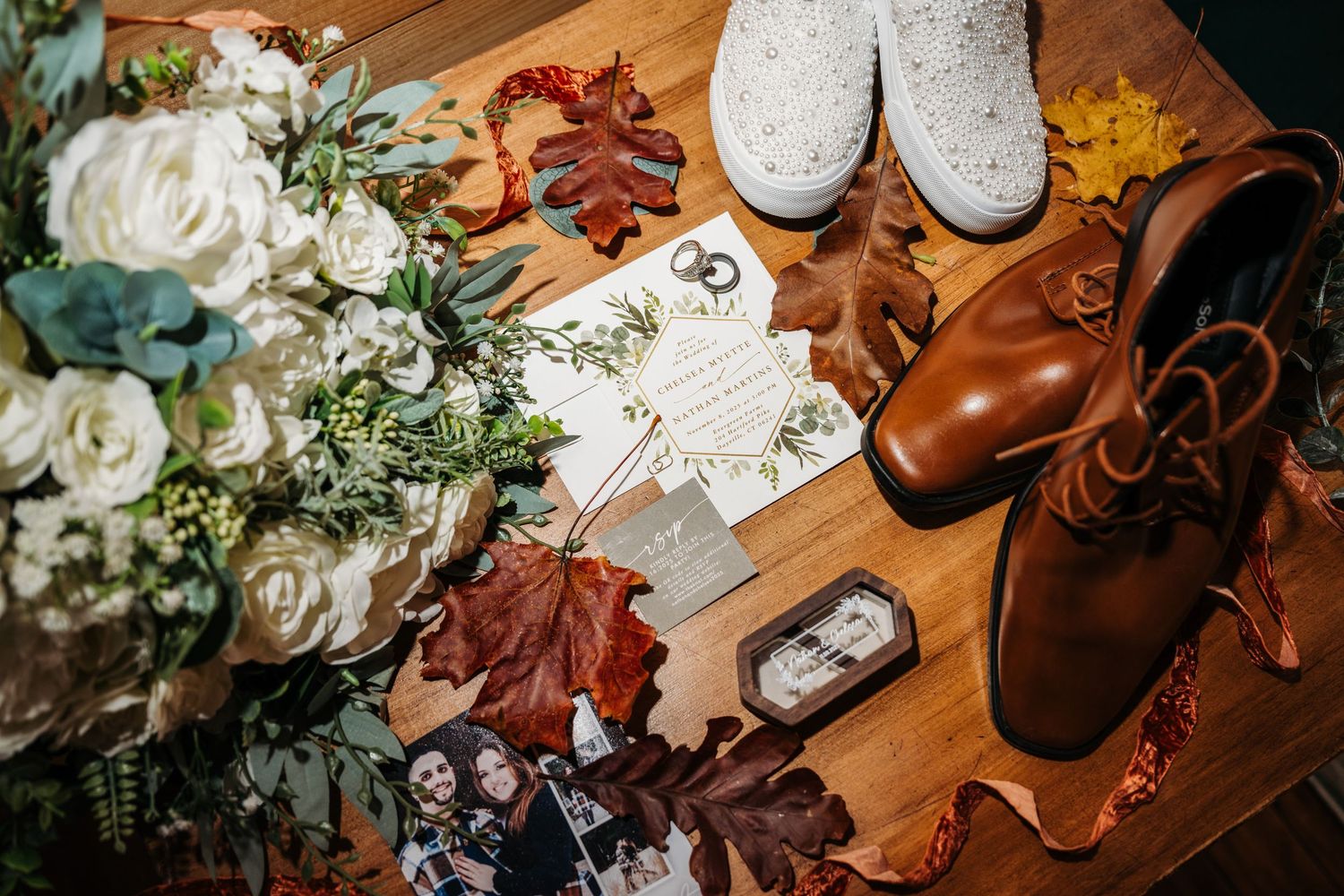 Fall wedding flat lay featuring white roses, brown leather shoes, invitation suite, and autumn leaves on wood.