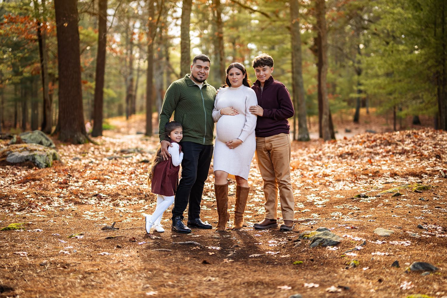 Family of four posing in autumn forest, pregnant mother in white dress surrounded by husband and two children.