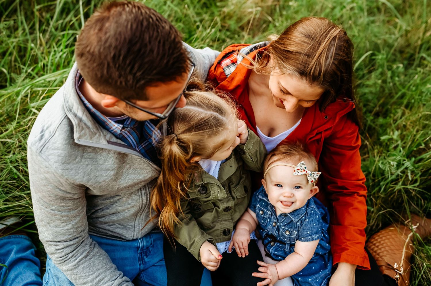 Family of four sitting in green grass, parents lovingly gazing at two young daughters during outdoor photo session.