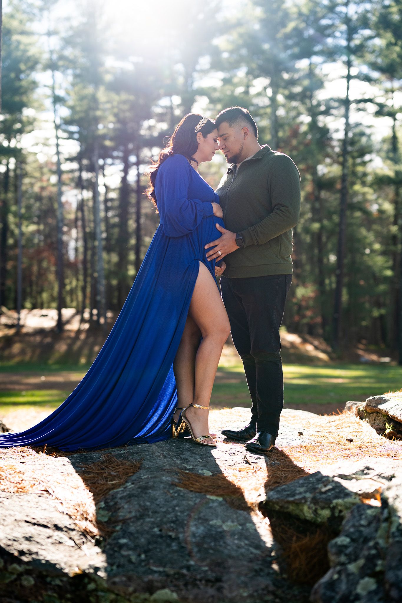 Expecting couple in tender embrace on rocks in sunlit pine forest during maternity photoshoot.