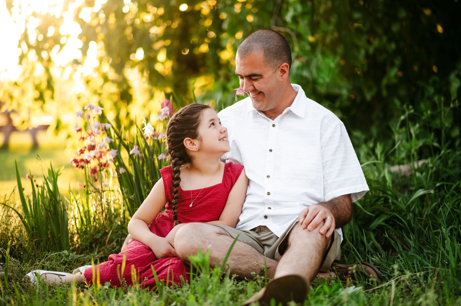 A father and daughter sit together in a lush green garden, sharing a warm smile in golden sunlight.