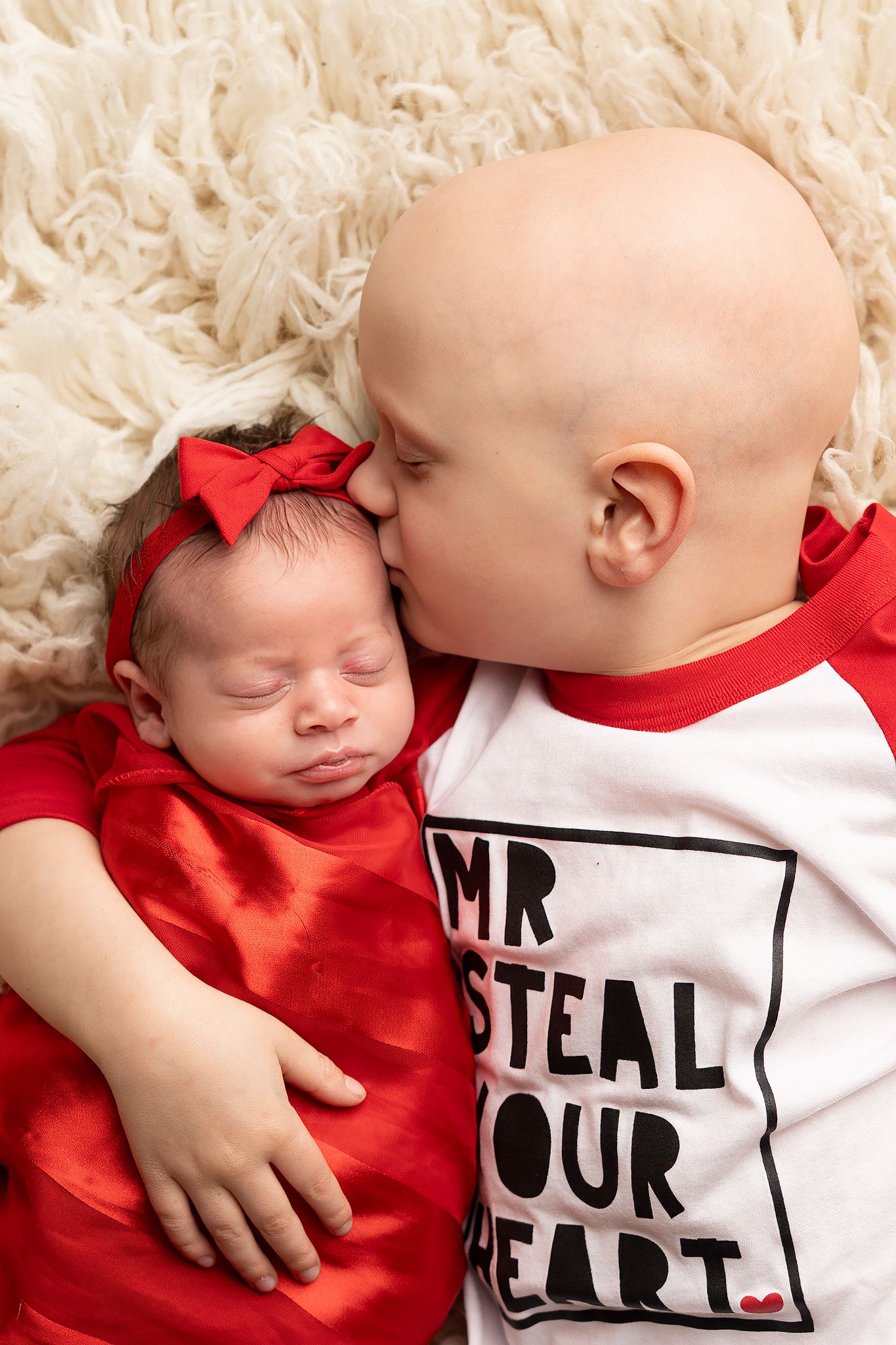 A young bald toddler in a Mr. Steal Your Heart shirt kisses a sleeping newborn baby girl in a red outfit with bow.