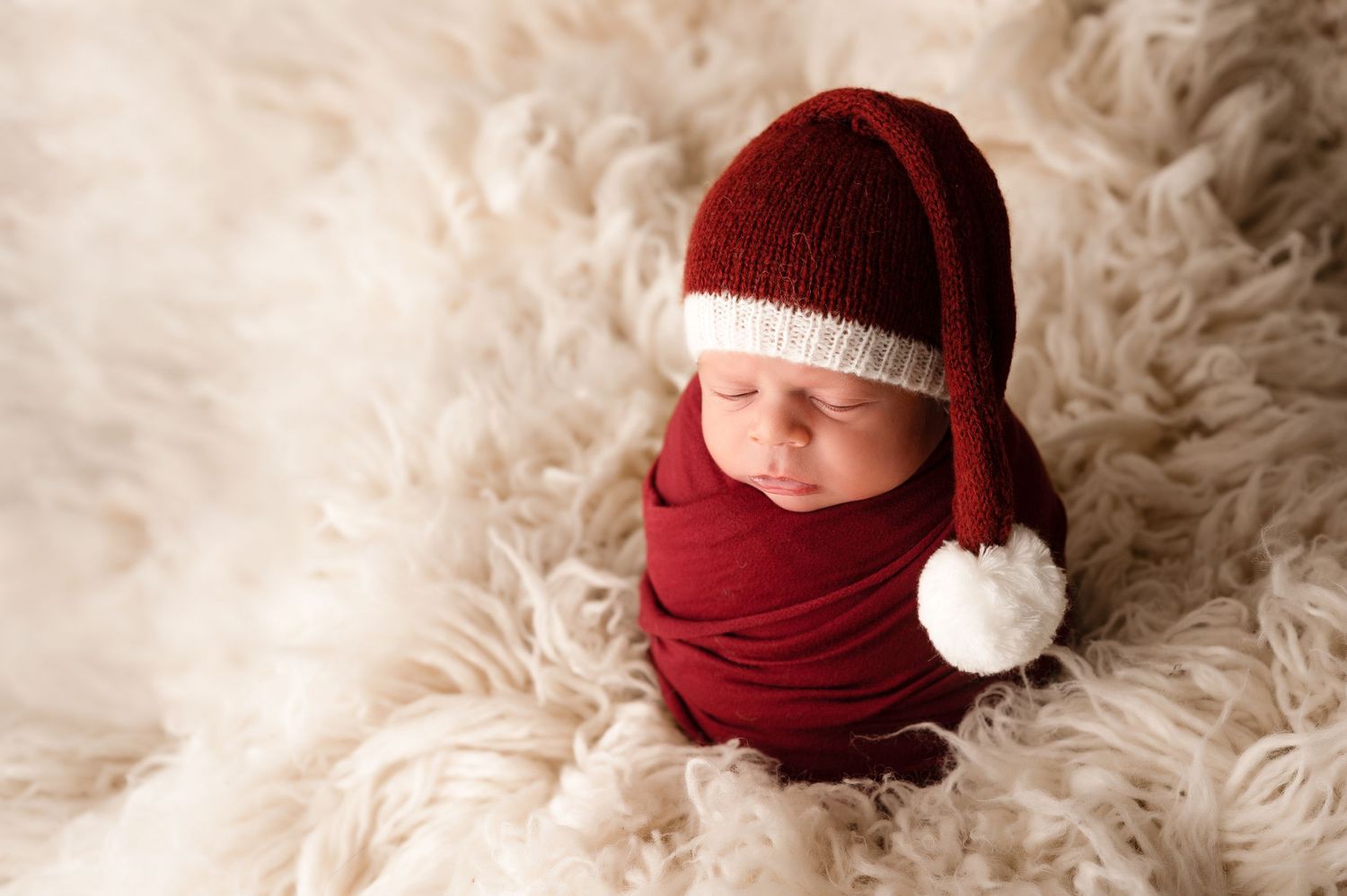 Newborn baby wrapped in red swaddle wearing a Santa hat on a fluffy cream rug for Christmas photo.
