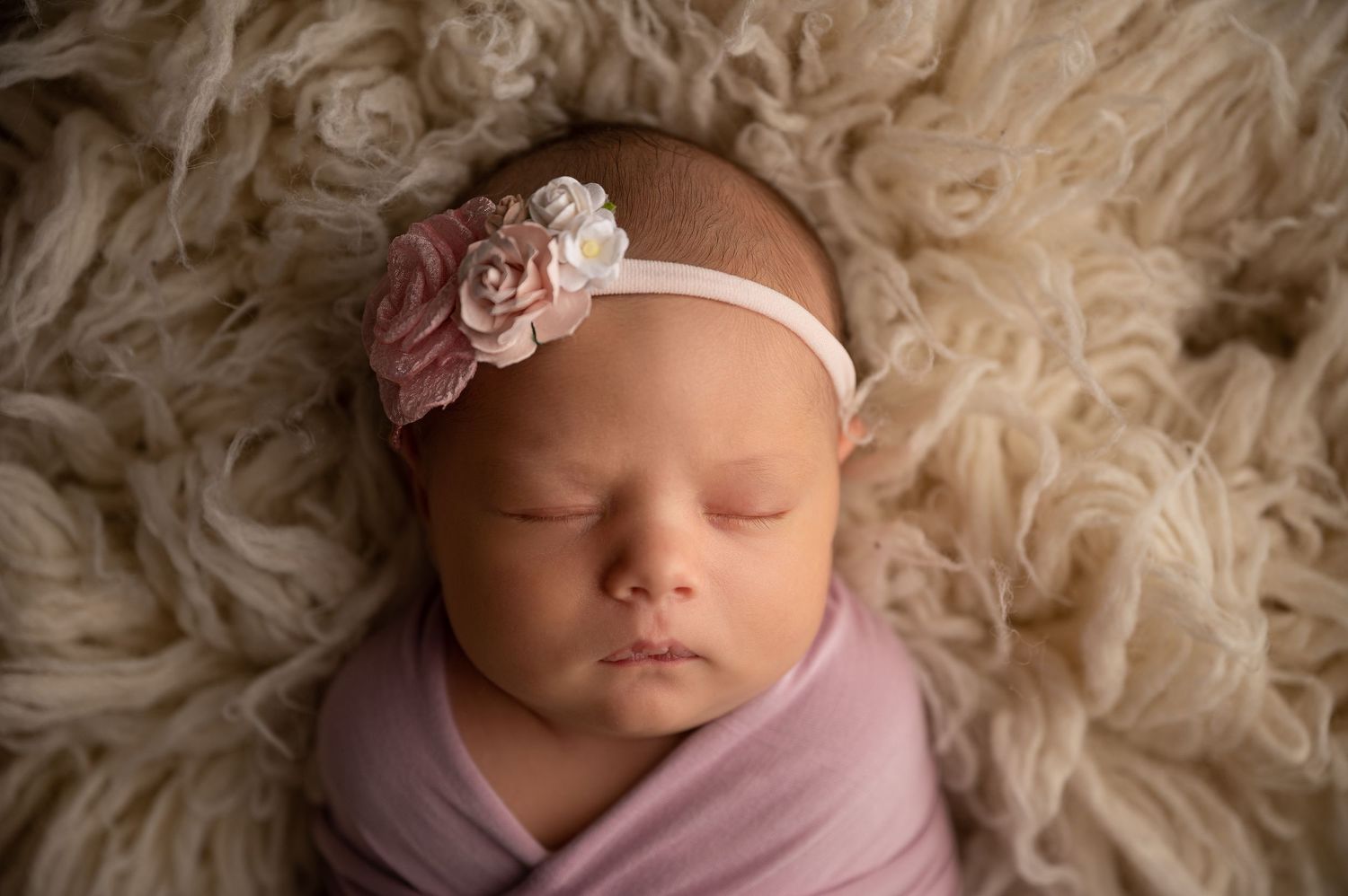 Sleeping newborn baby girl wrapped in pink swaddle with floral headband on fluffy cream backdrop.