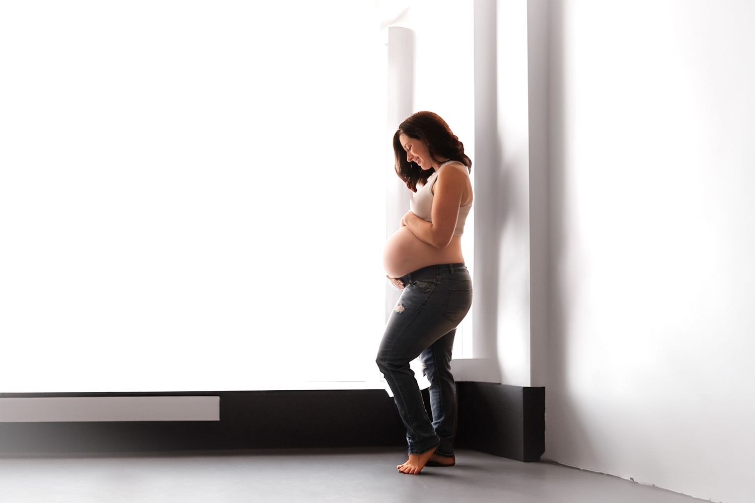 Pregnant woman in jeans cradling her baby bump while leaning against a bright white wall barefoot.