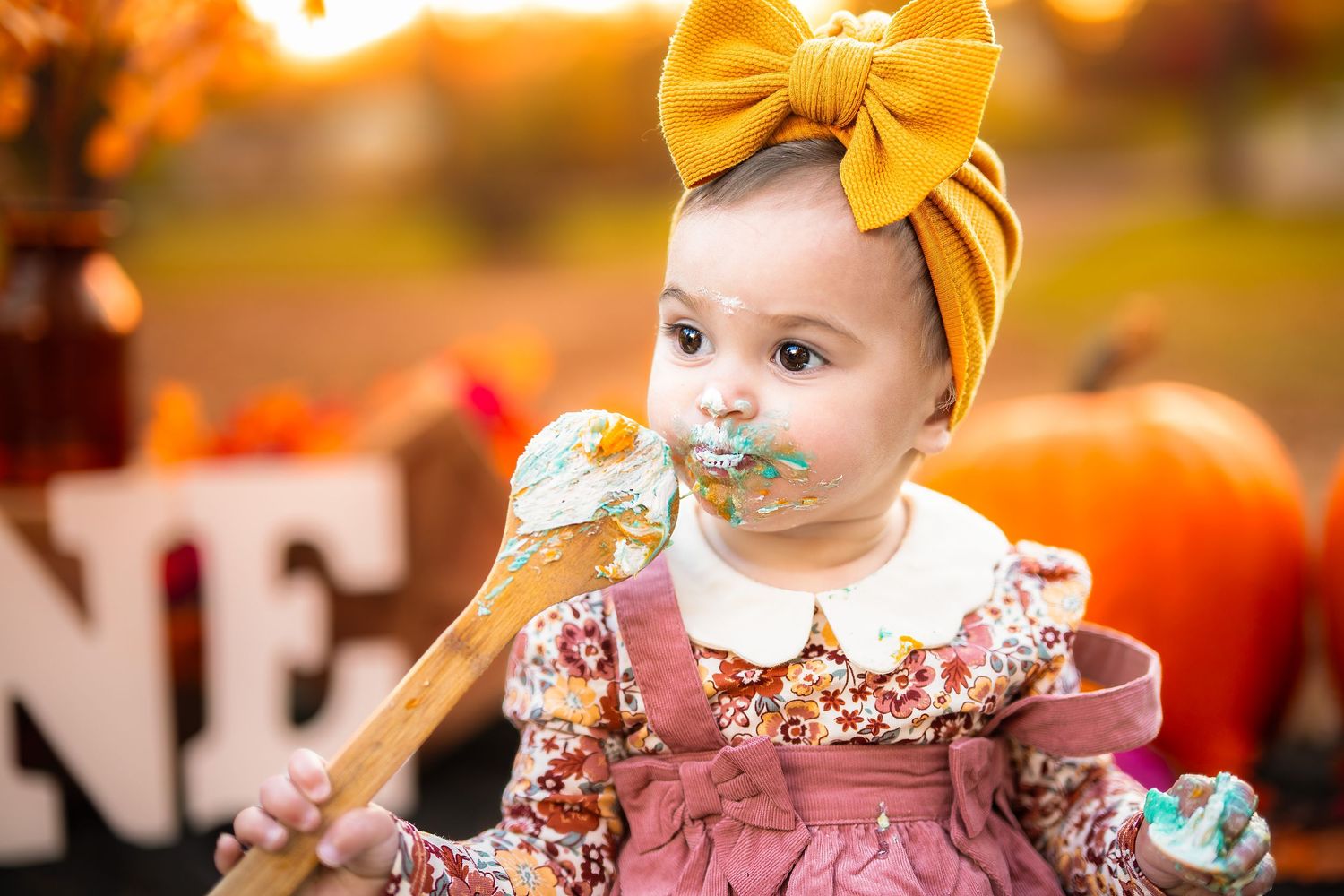 Adorable baby girl in floral dress and mustard bow headband eating cake at a fall-themed first birthday photoshoot.