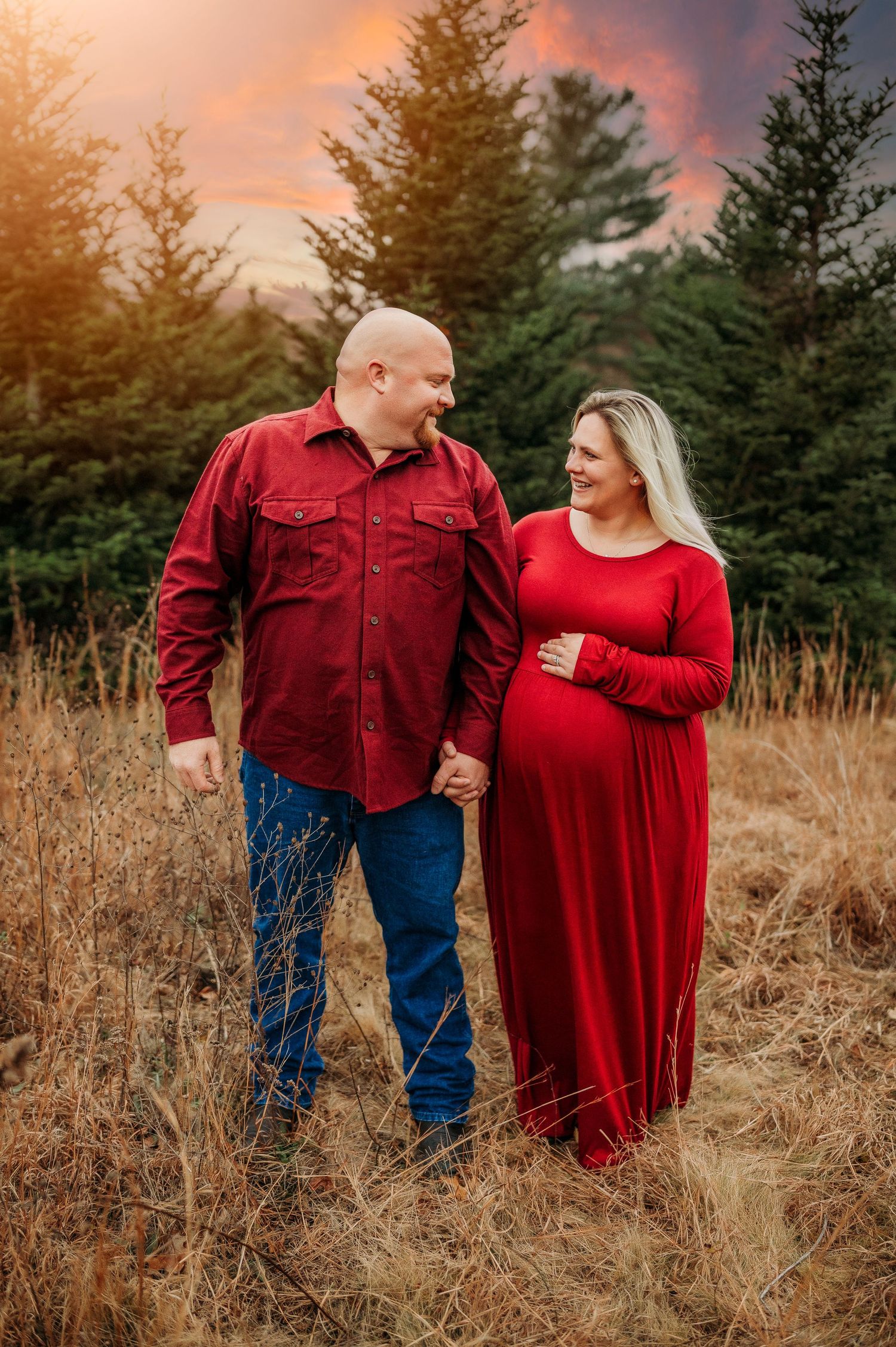 Pregnant couple in red holding hands in a golden field at sunset with evergreen trees and dramatic sky behind them.