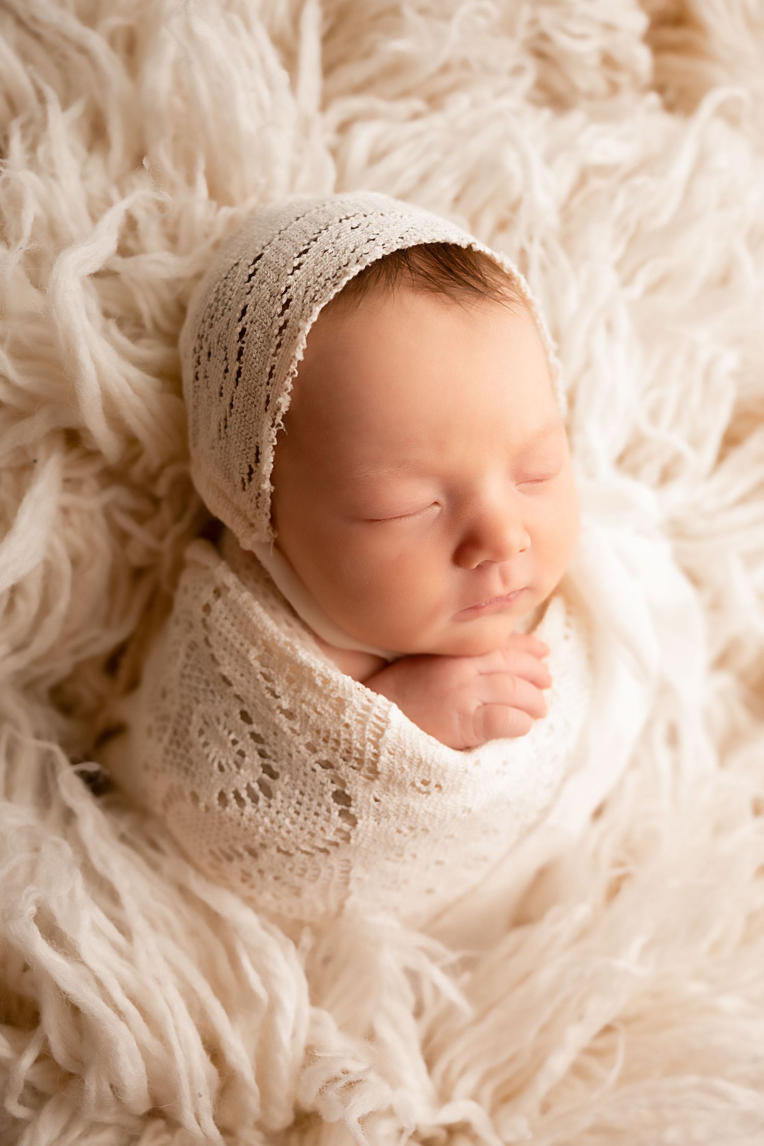 Sleeping newborn baby wrapped in cream knit blanket and bonnet on fluffy white fur backdrop.