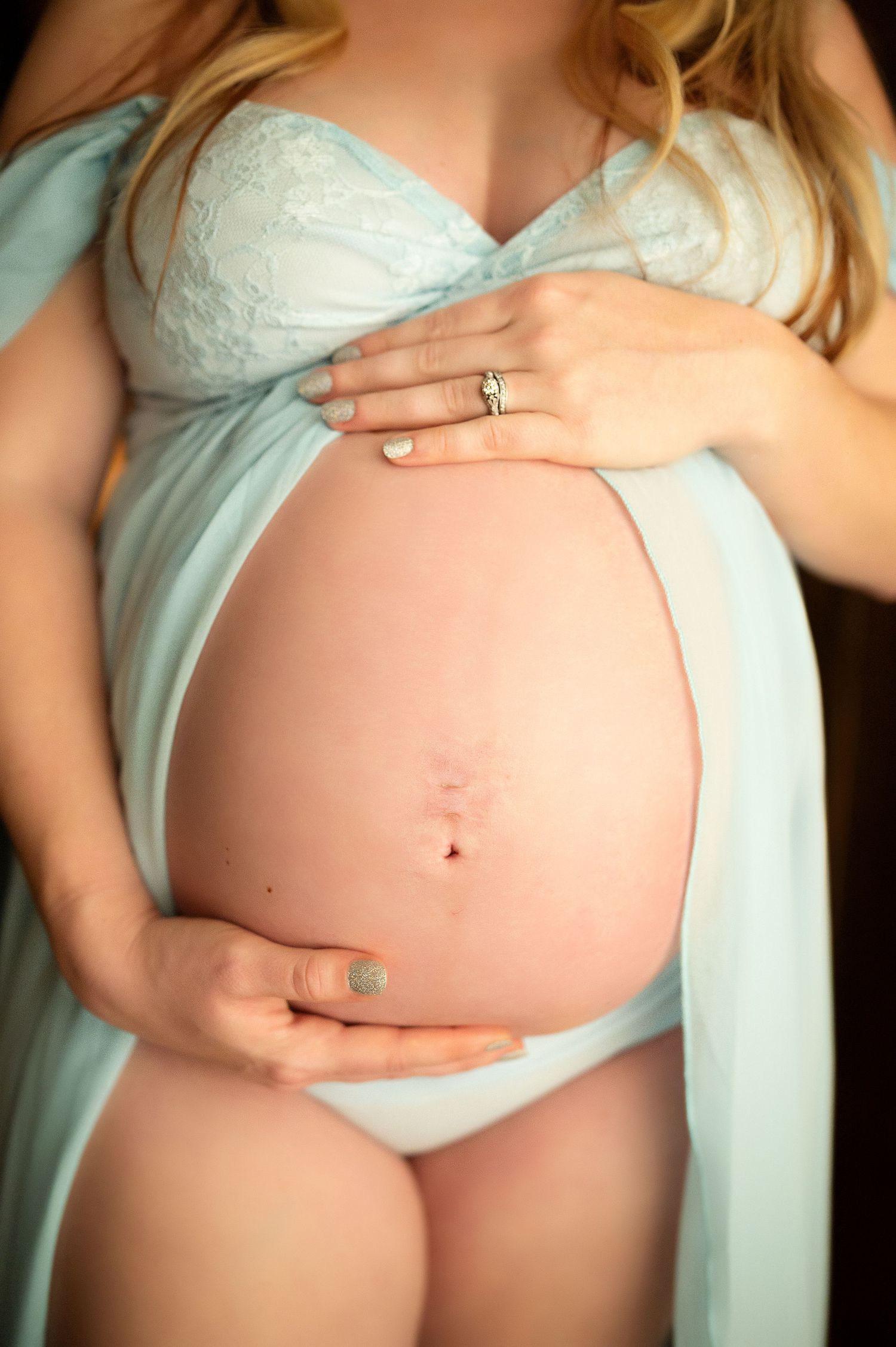 Pregnant woman in light blue lace lingerie cradling her bare baby bump, wearing an engagement ring.