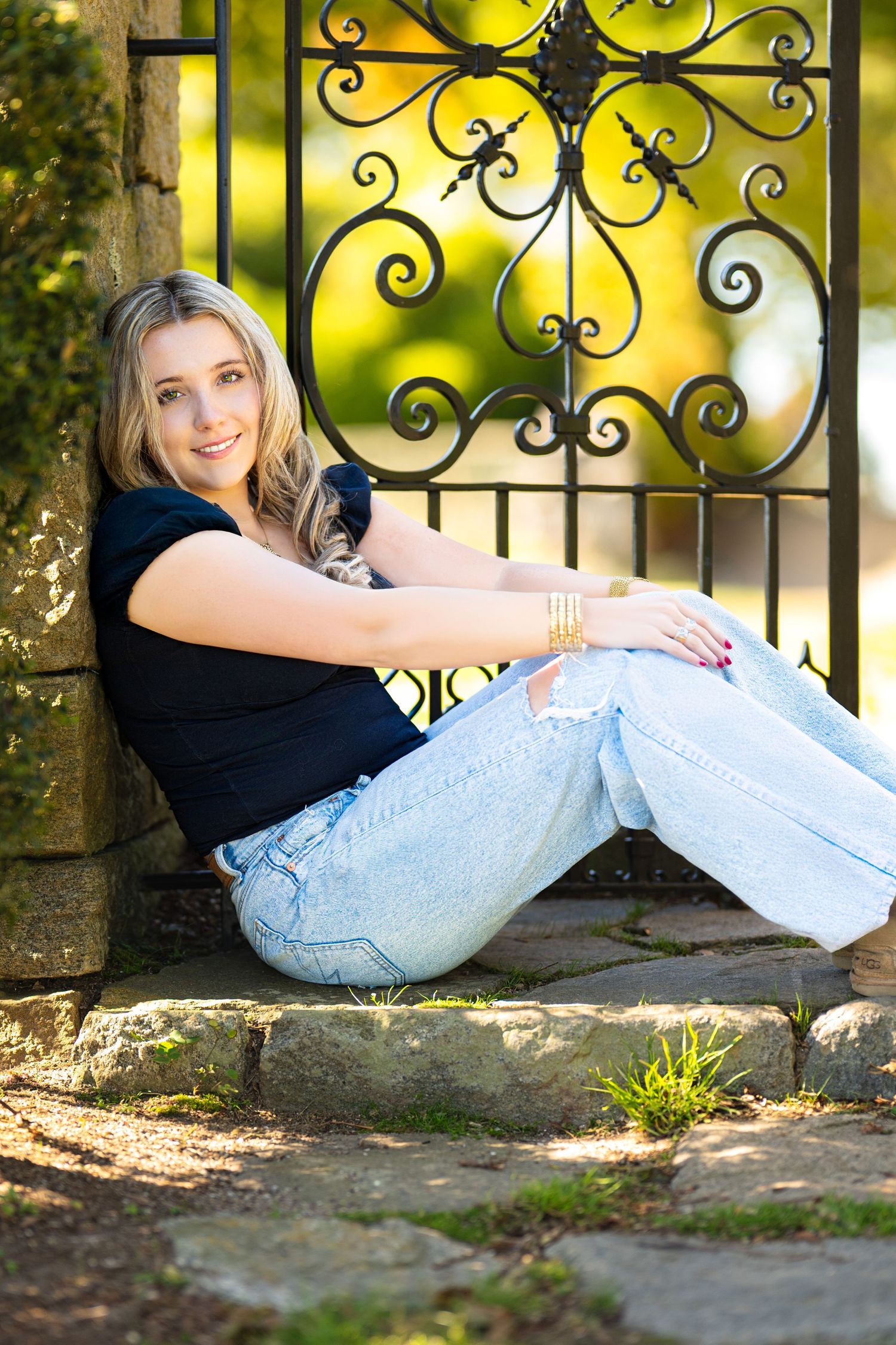 Young blonde woman in black top and ripped jeans sitting against ornate iron gate on stone steps outdoors.