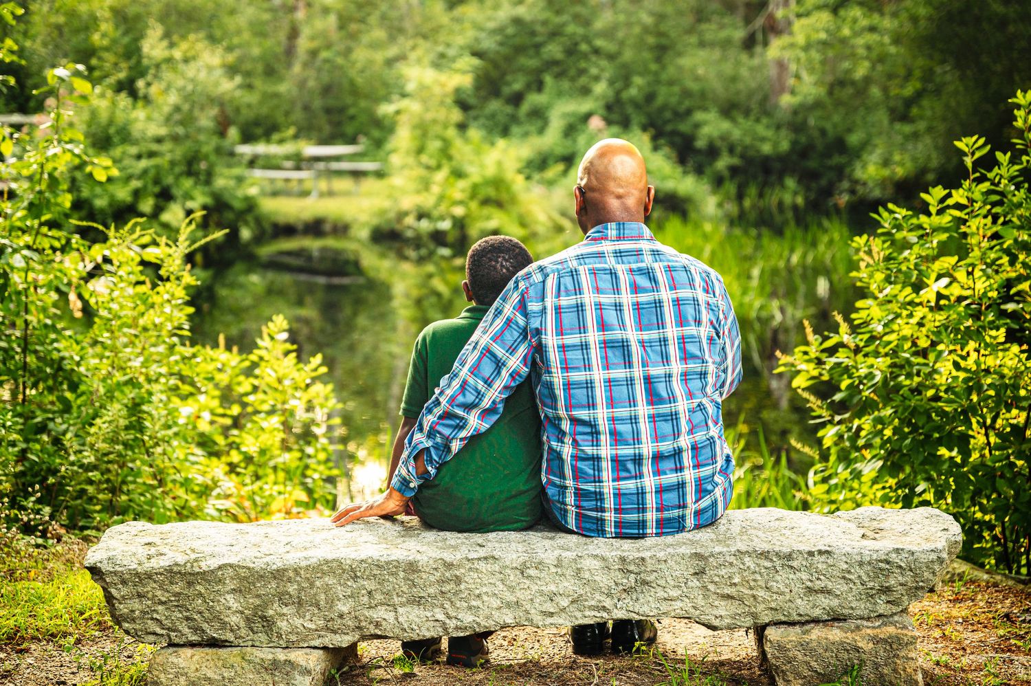 Adult and child sitting together on a stone bench overlooking a serene pond surrounded by lush green trees.