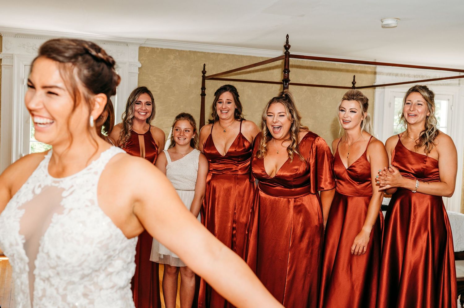 Bride in white lace gown laughs as bridesmaids in rust satin dresses react with joy in elegant bedroom.