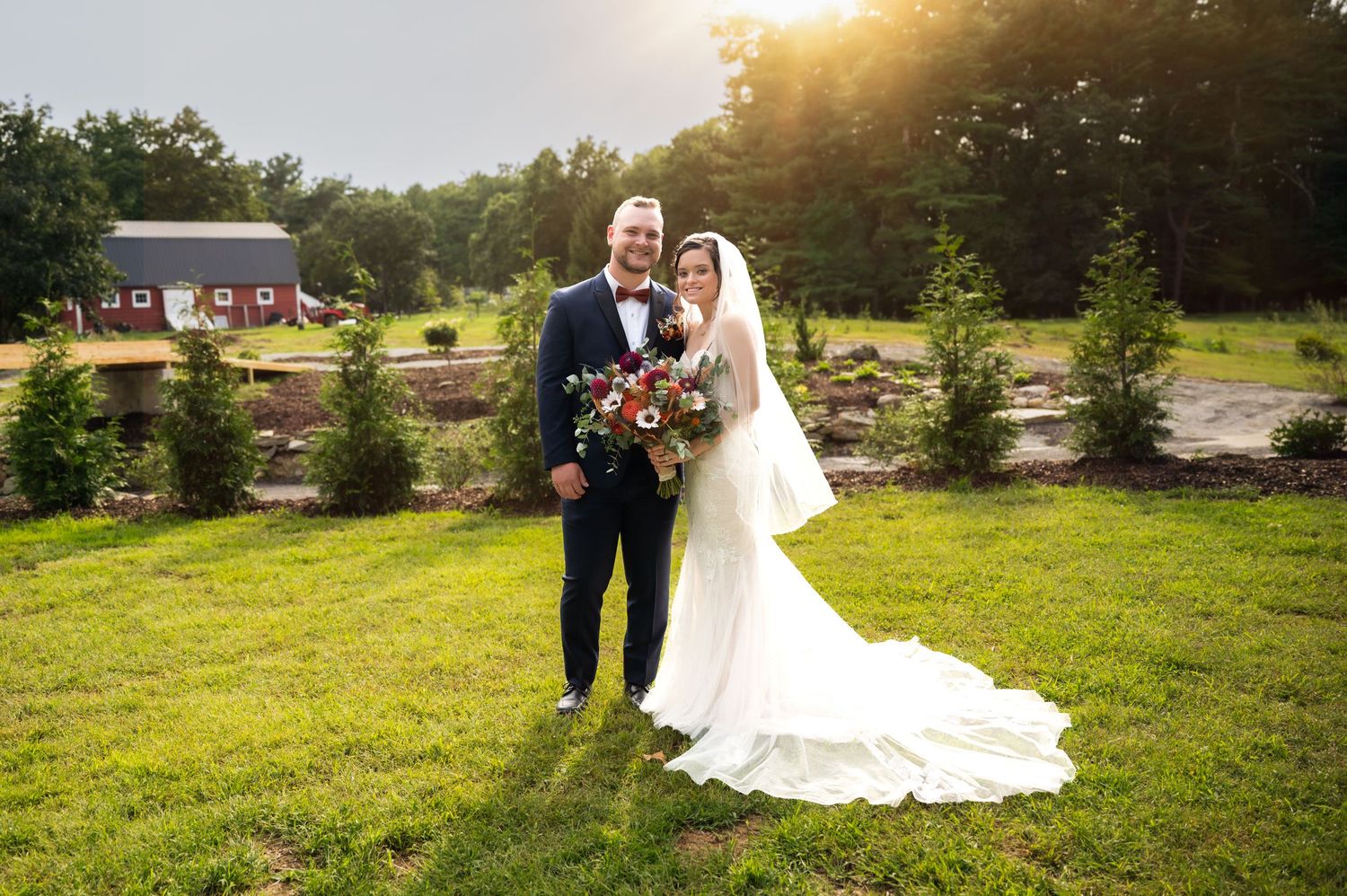 Bride and groom posing together on lush green lawn at golden hour with red barn and trees in background.