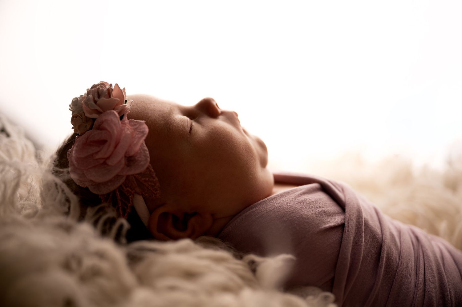 Sleeping newborn baby girl wrapped in pink swaddle with floral headband on fluffy white blanket.