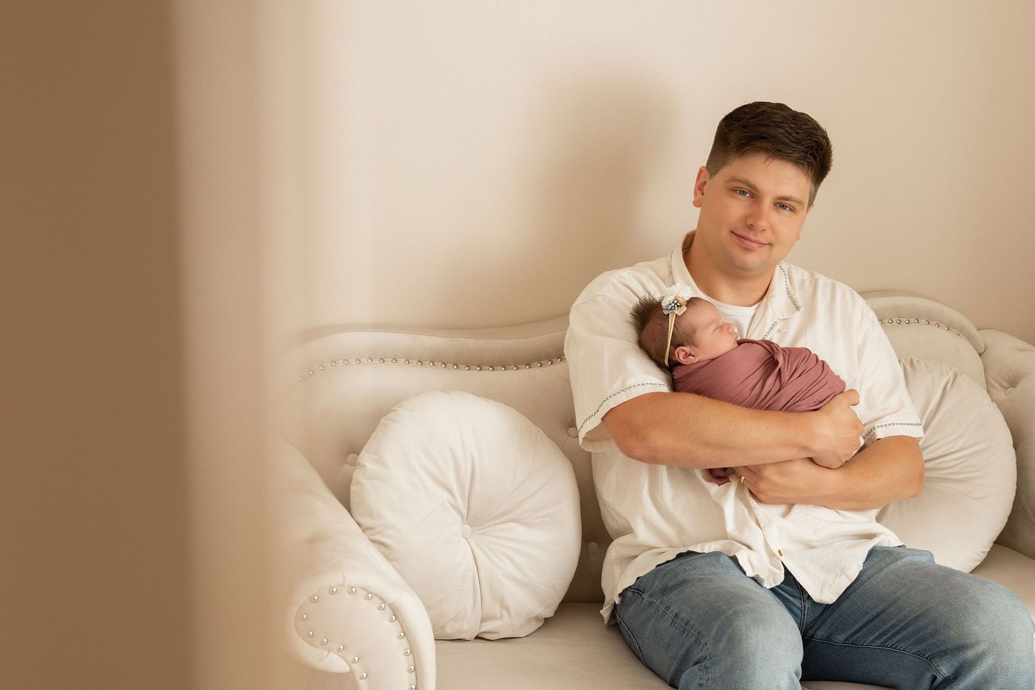 Young father sitting on a white couch, gently cradling his sleeping newborn baby wrapped in a pink blanket.Father gently holding his newborn