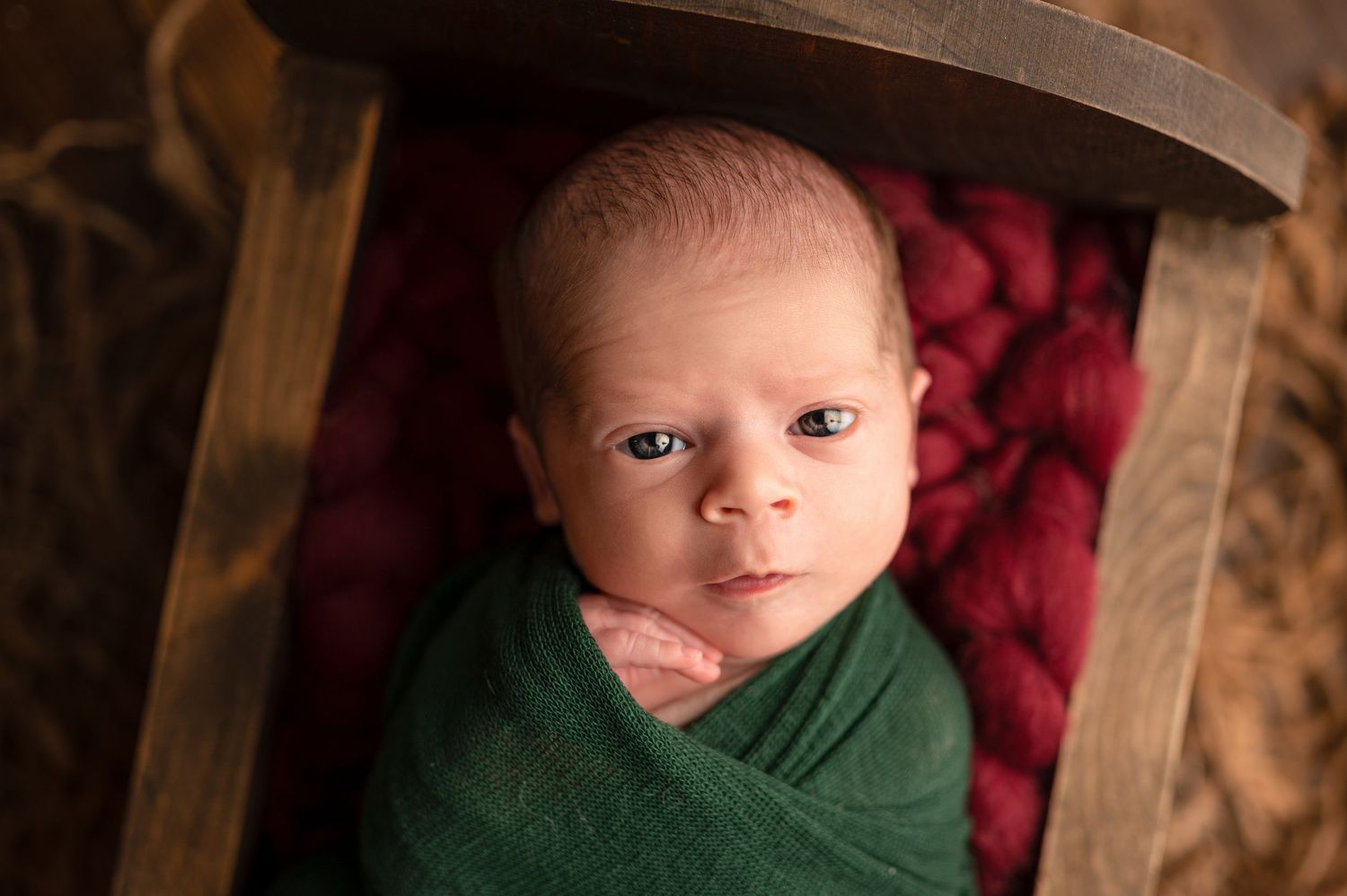 Newborn baby swaddled in green wrap resting in wooden bowl with red knit blanket, professional newborn photography.