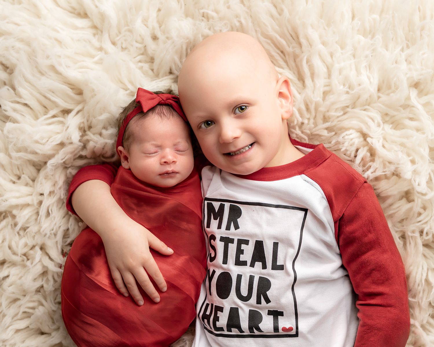 Toddler boy in Mr. Steal Your Heart shirt cuddles sleeping newborn baby girl in red swaddle on fluffy rug.