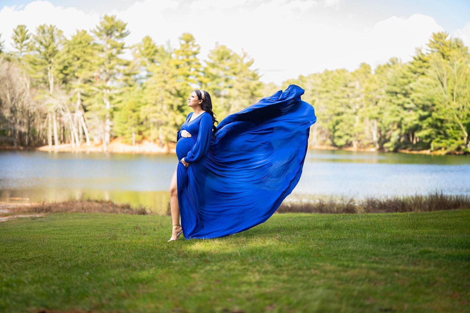 Pregnant woman in a flowing royal blue gown posing outdoors by a lake with trees.