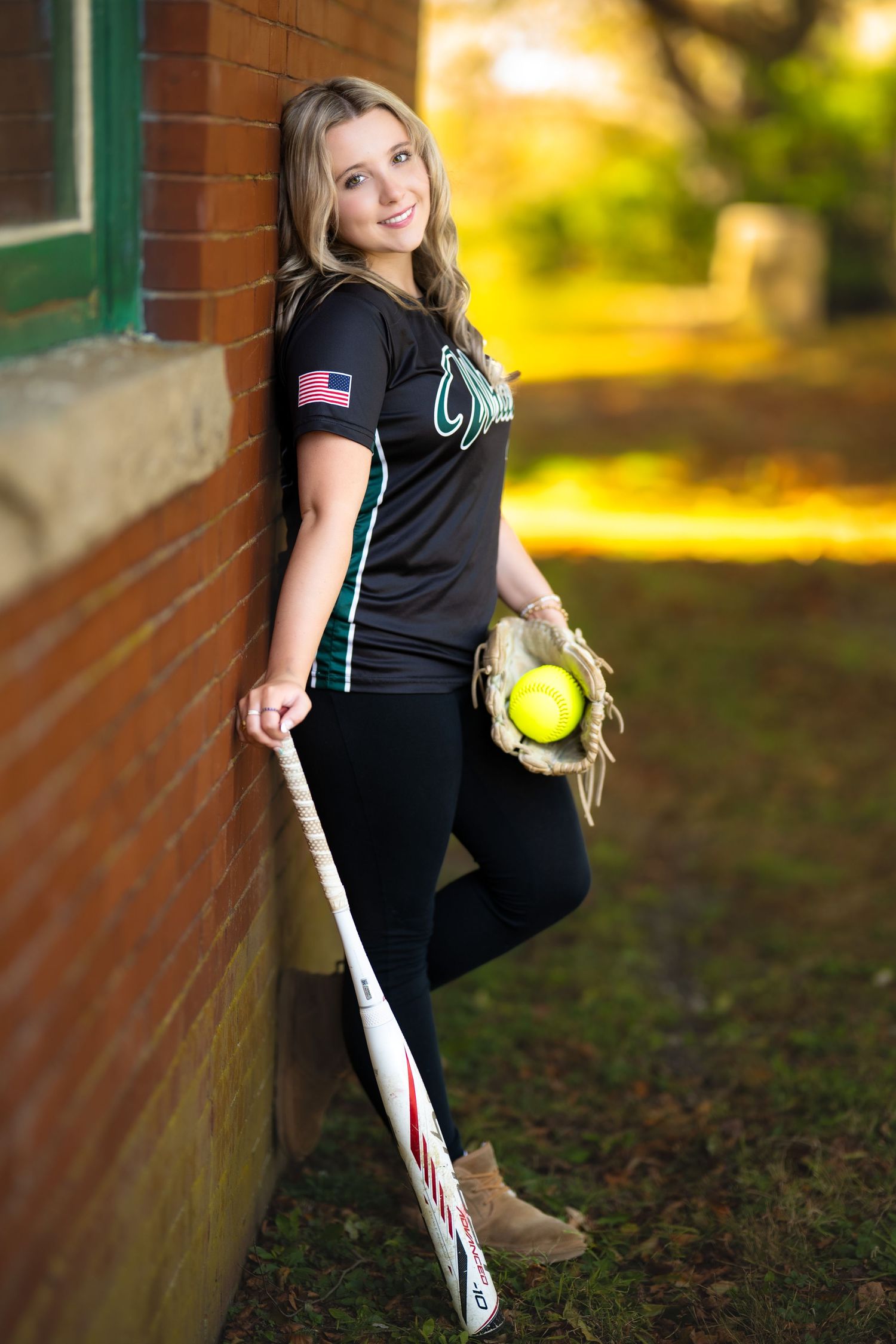 Teen girl in black softball jersey leaning on brick wall holding bat and glove with yellow softball.
