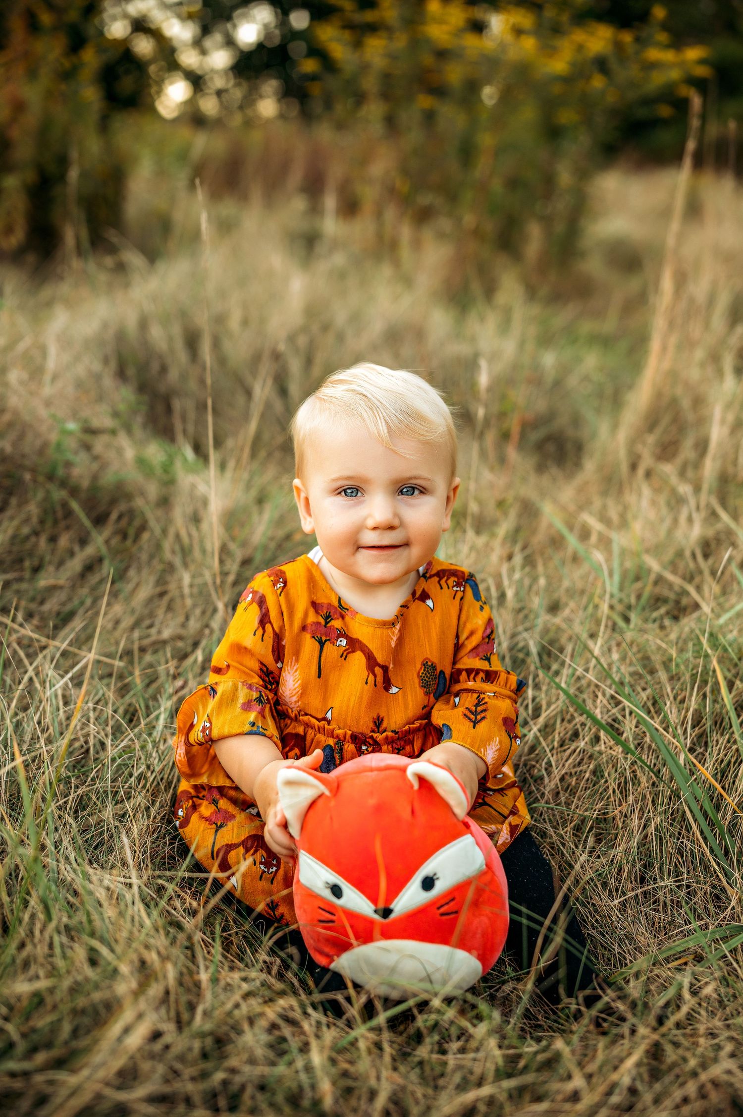 Toddler in orange fox dress sits in grass field holding a red fox stuffed animal toy outdoors in autumn.