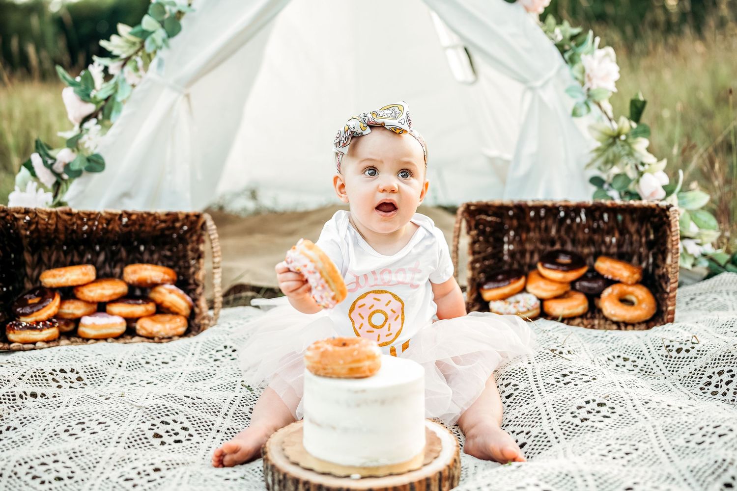 Baby girl in crown celebrates first birthday with white cake and donuts on blanket outdoors near teepee.