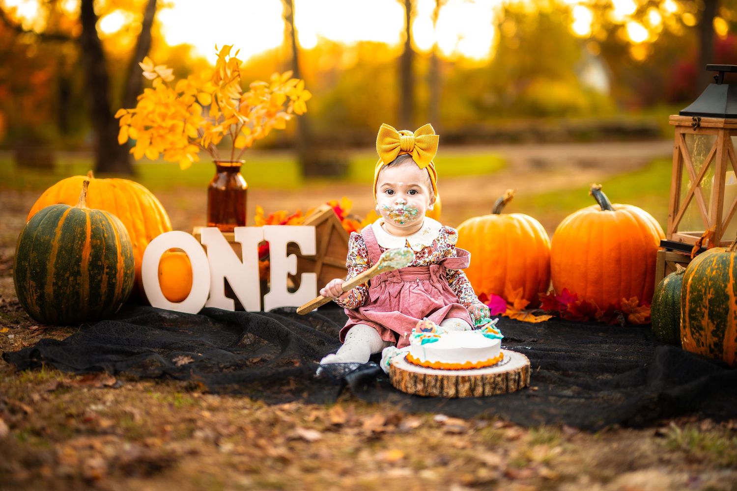 Baby girl in pink dress celebrates first birthday with pumpkins and ONE sign at fall outdoor cake smash photo session.