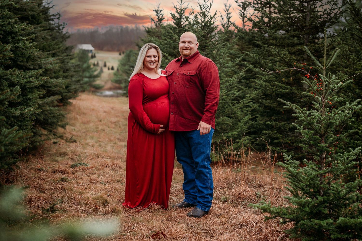 Pregnant couple in red outfits posing at a Christmas tree farm during a maternity photo shoot at sunset.